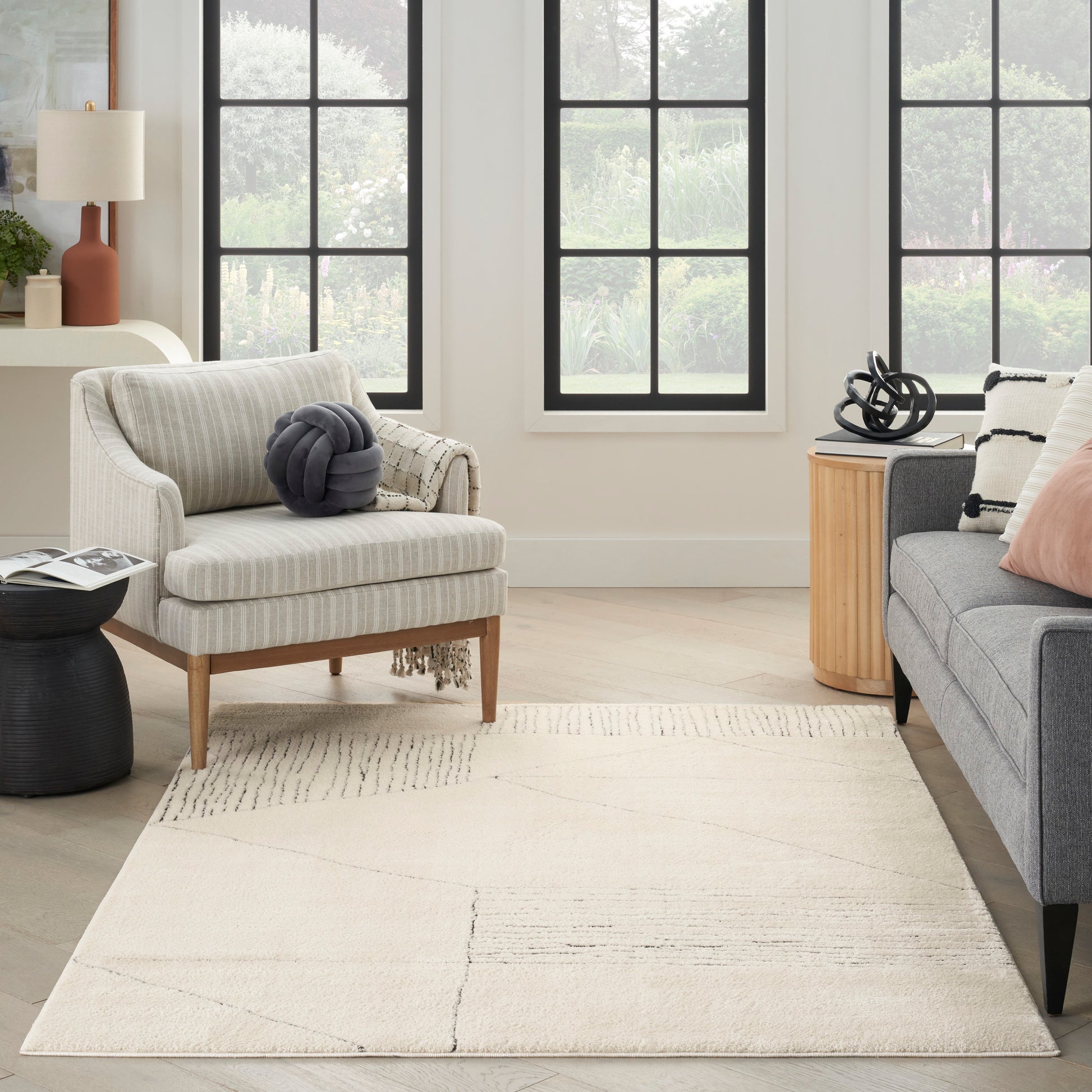 Beige striped armchair with wooden legs and a gray knot pillow beside a gray sofa; cream geometric rug and round wood side table.