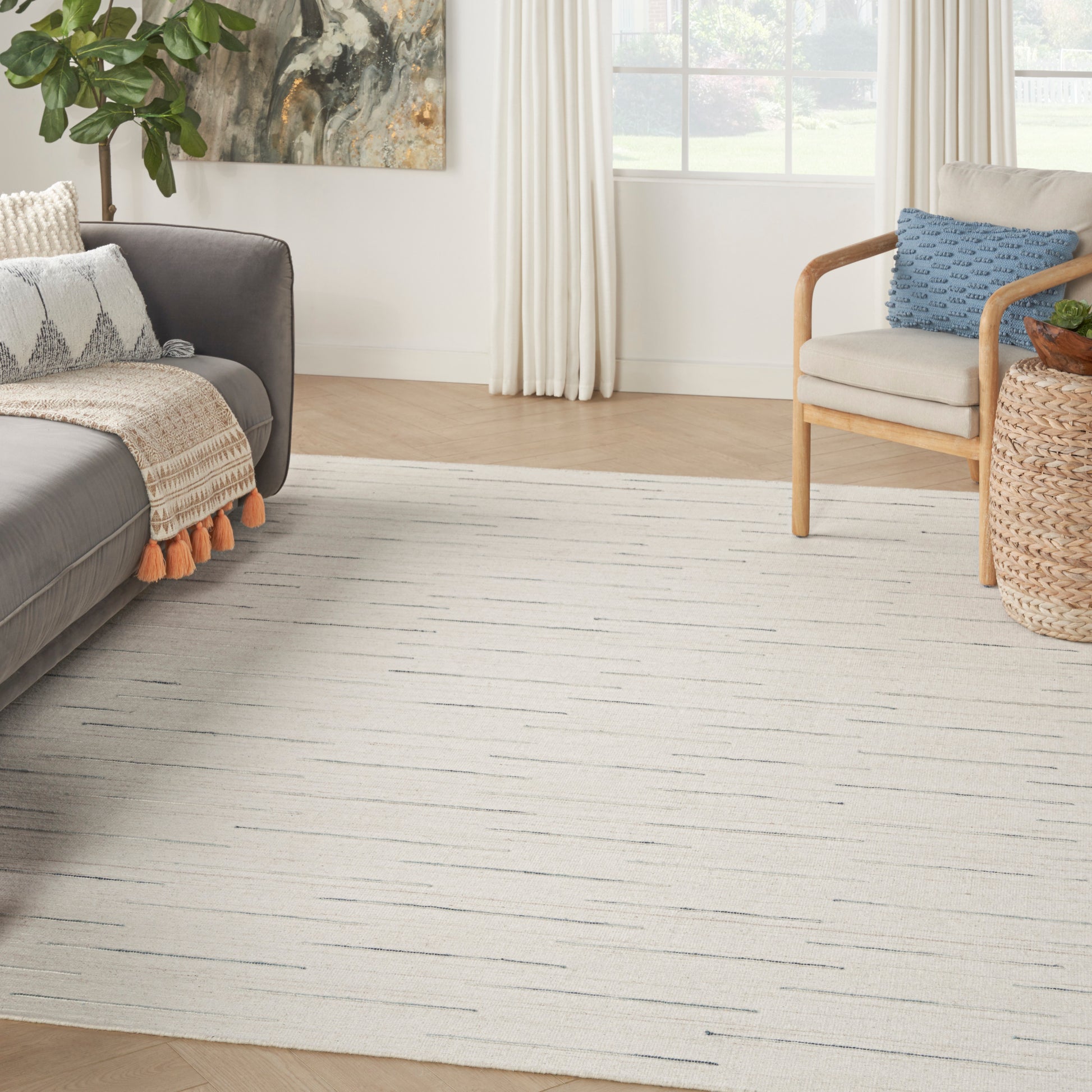 Cream woven area rug with subtle gray linear pattern in a bright living room with a gray sofa, light wood chair, and wicker side table.