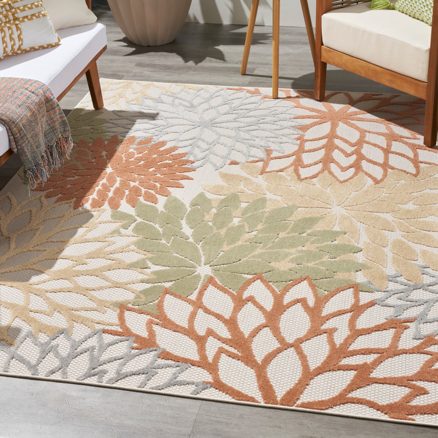 Outdoor woven area rug with large leaf motifs in orange, sage green, beige and gray; laid on a sunlit patio beside white seating.