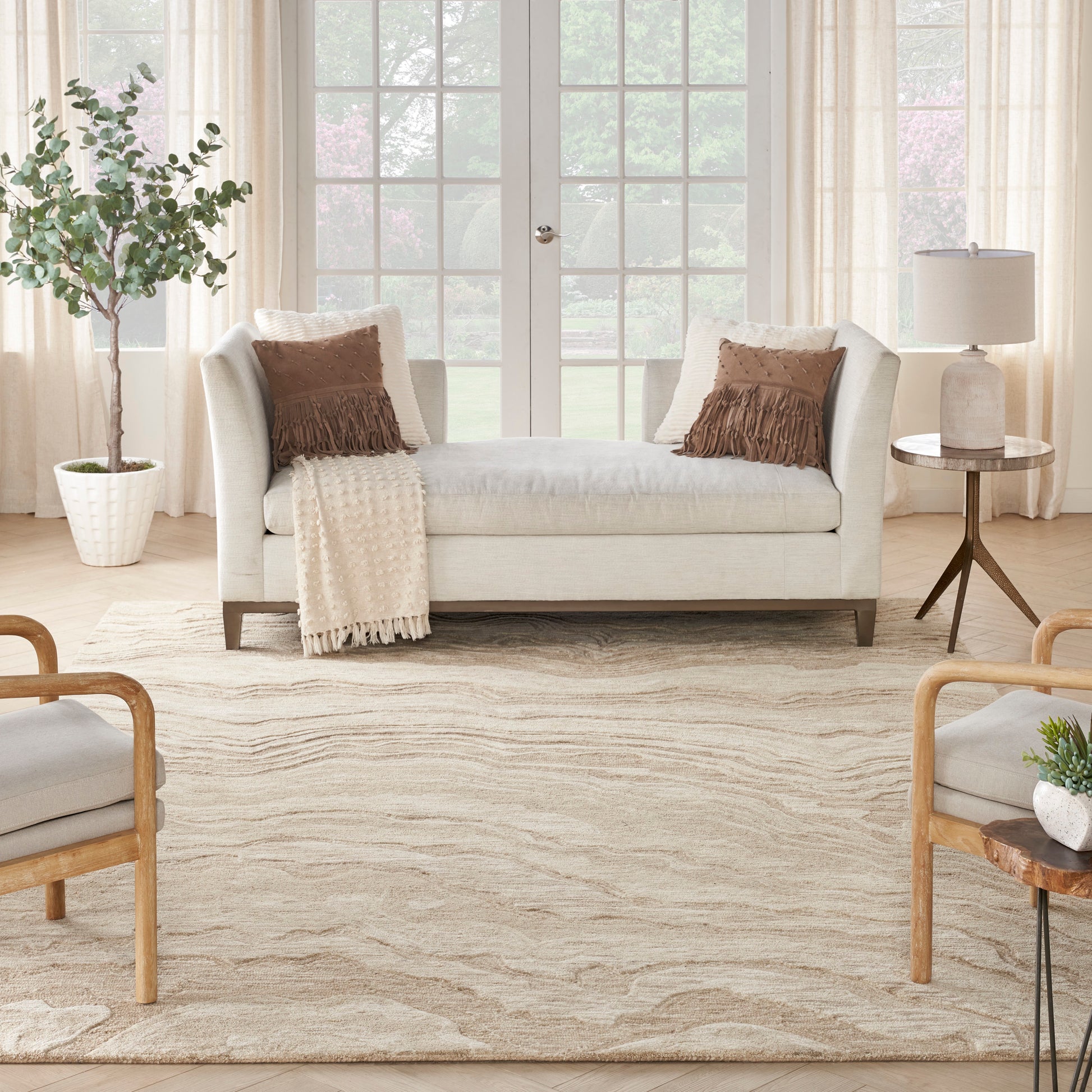Cream rug in a bright living room, paired with a beige sofa and brown fringe pillows, a beige throw, a potted plant, and a round side table with a lamp.