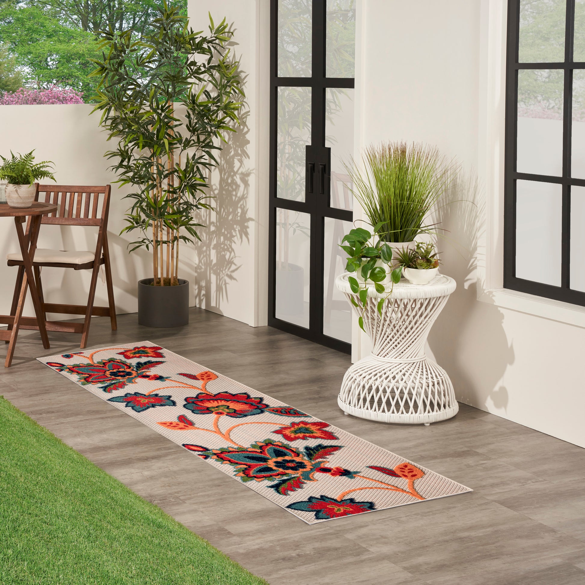 Long floral runner rug on wood floor by glass doors; potted plants, white wicker side table, and garden seating.