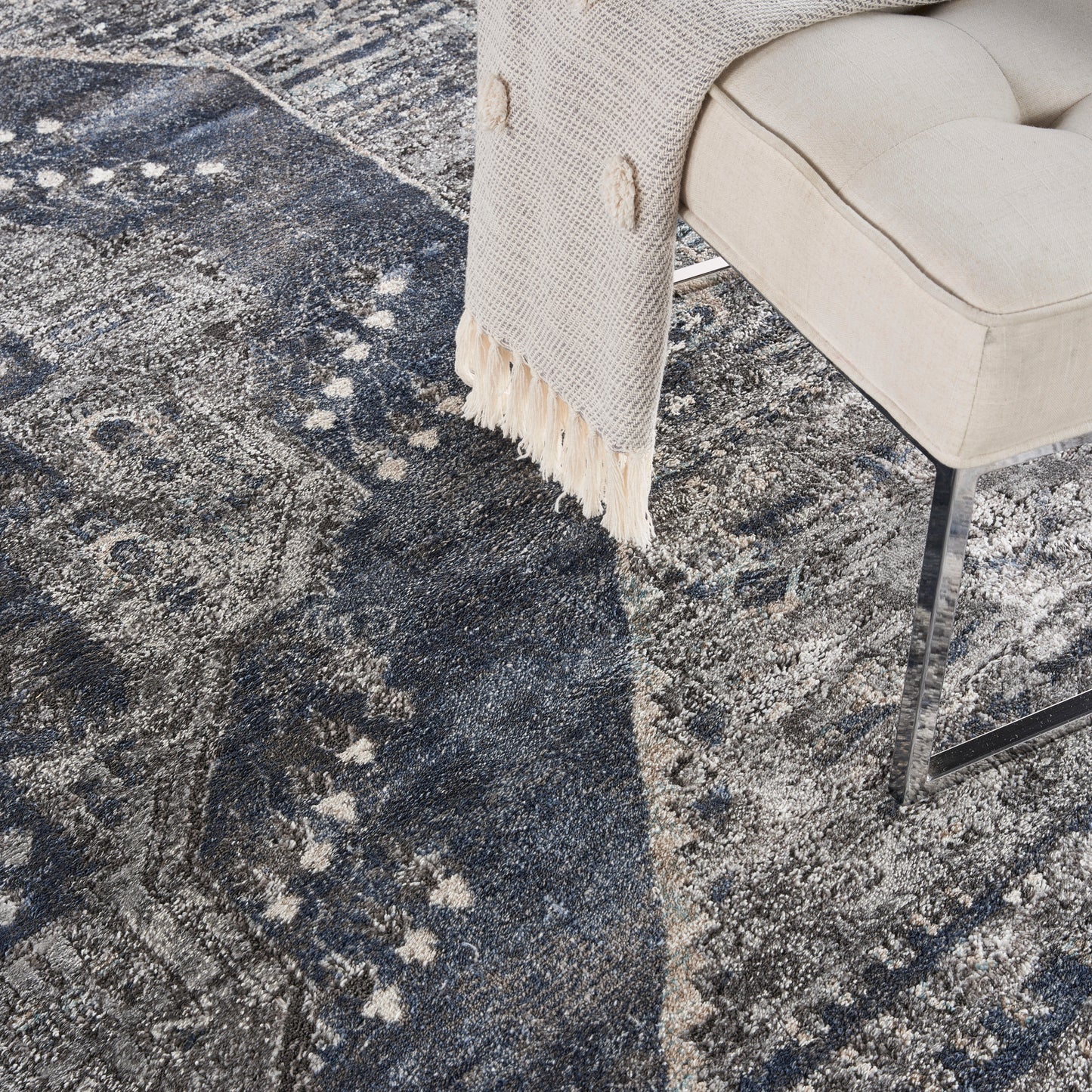 Beige upholstered chair with metal legs, draped cream fringe throw, on a dark blue abstract patterned rug.