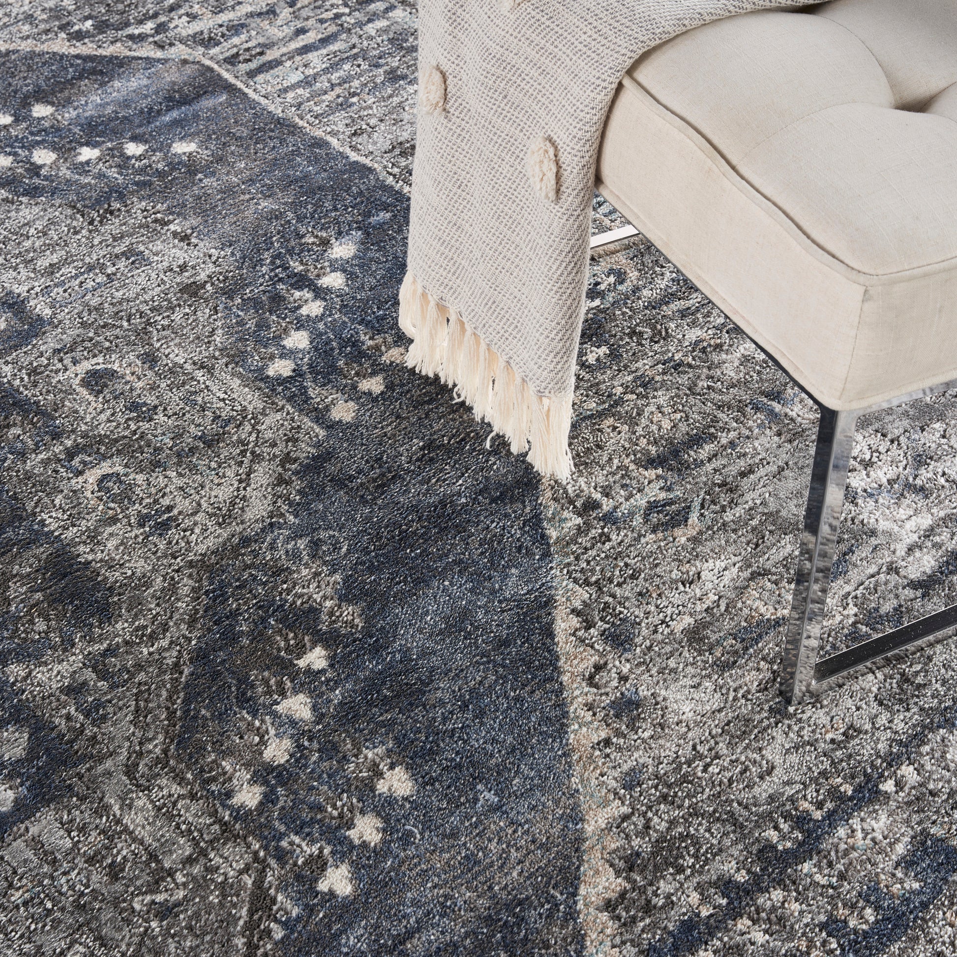 Beige upholstered chair with metal legs, draped cream fringe throw, on a dark blue abstract patterned rug.