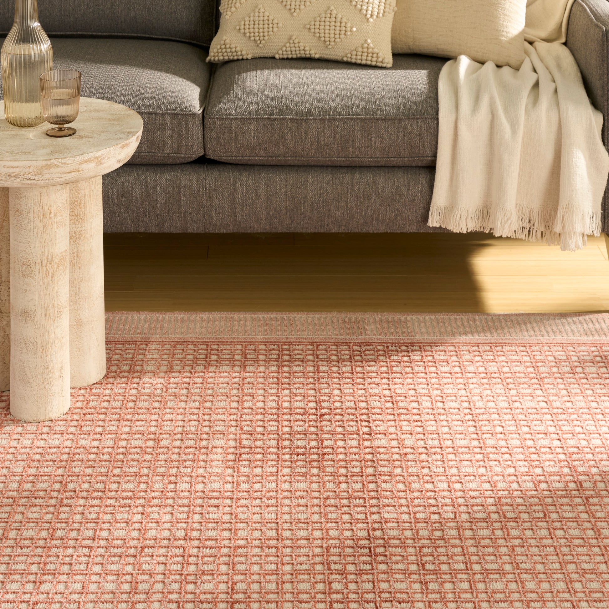 Cozy living room scene: pink geometric rug, round light wood side table, gray sofa, beige throw, knit cushion.