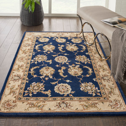 Rectangular rug with navy blue center and cream floral borders; beige border with scrolling vines on a wood floor.