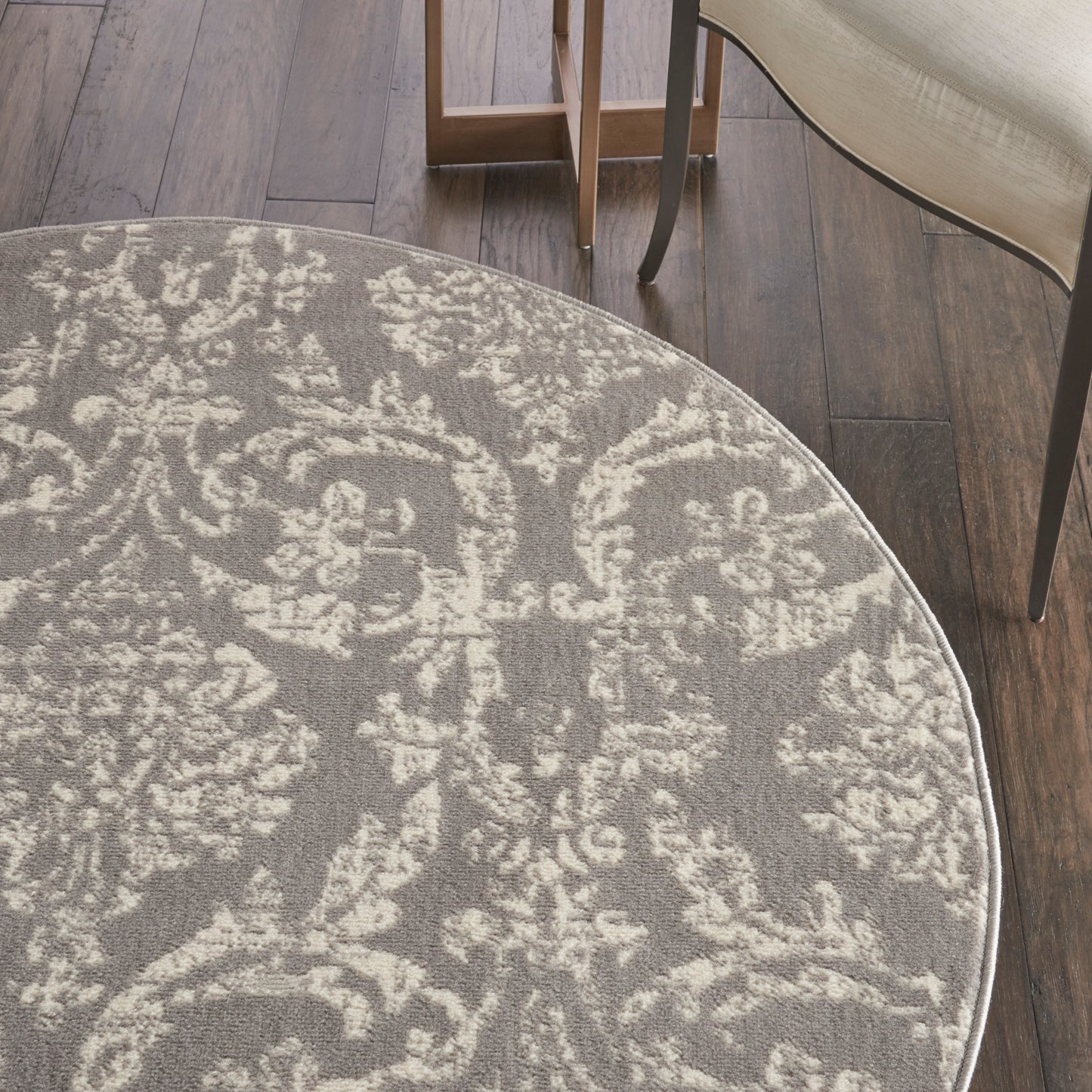 Round gray rug with ivory damask floral pattern on dark wood floor; beige upholstered chair legs visible.