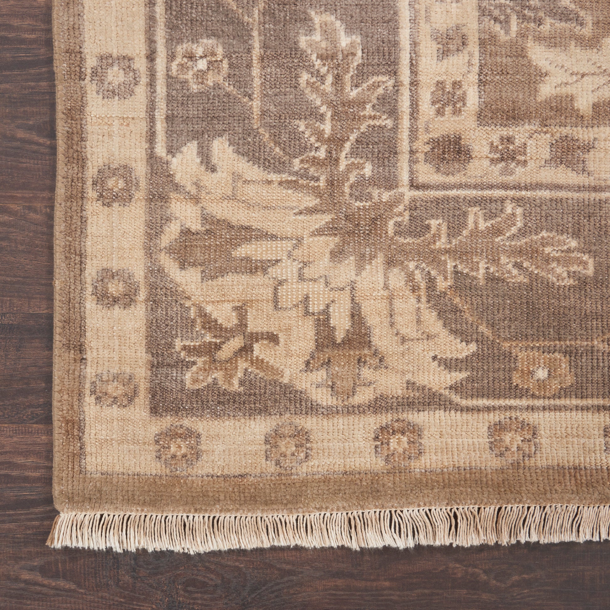 Beige-brown ornate area rug with floral motifs and fringe along the edge on a dark wood floor.