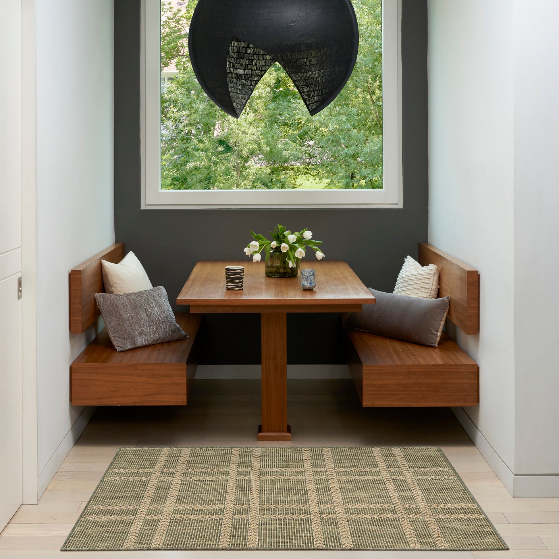 Wooden corner dining set with square table, benches, gray cushions, vase of flowers, black pendant lamp, window view.
