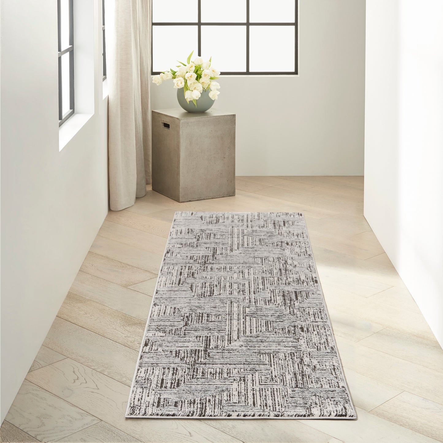 Grey geometric runner rug on pale wood floor in bright hallway with concrete pedestal and white flowers by the window.