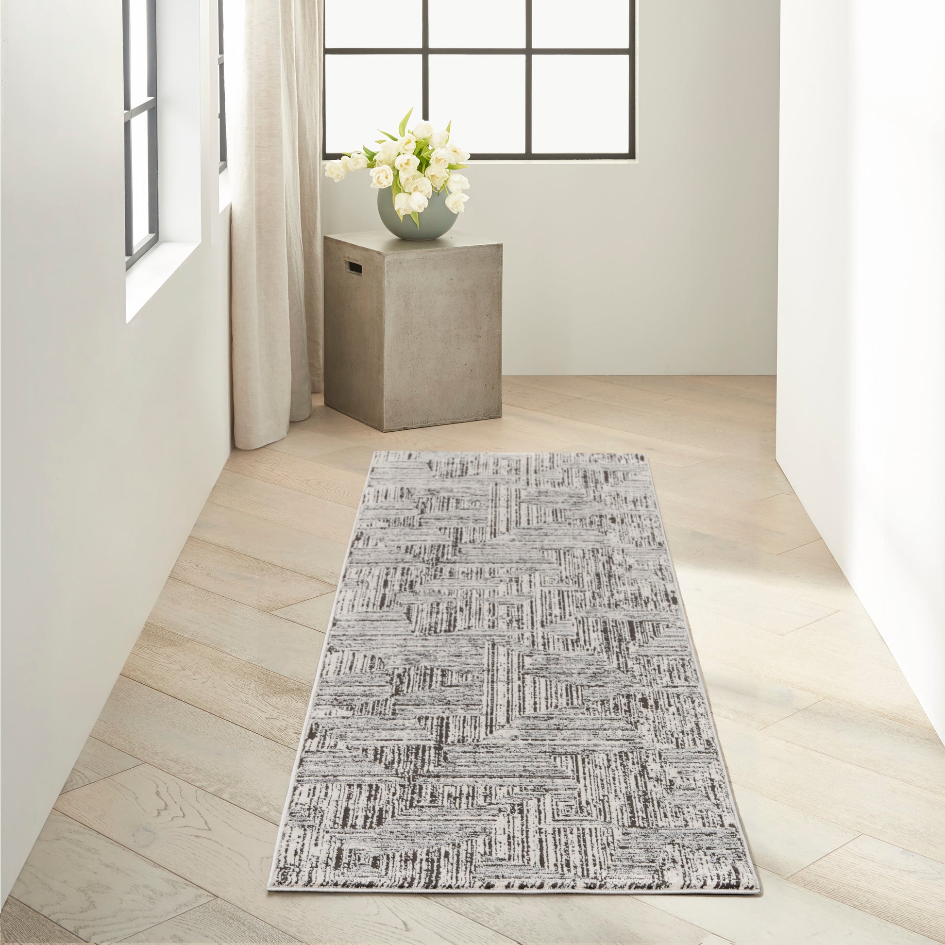 Grey geometric runner rug on pale wood floor in bright hallway with concrete pedestal and white flowers by the window.