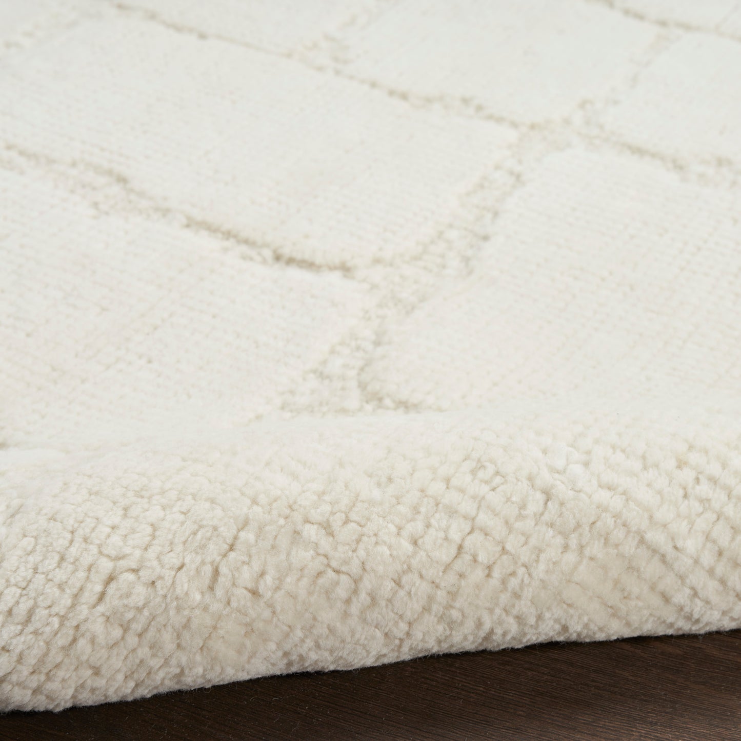 Cream plush rug close-up with thick pile and subtle grid pattern; edge folded on dark wood floor.