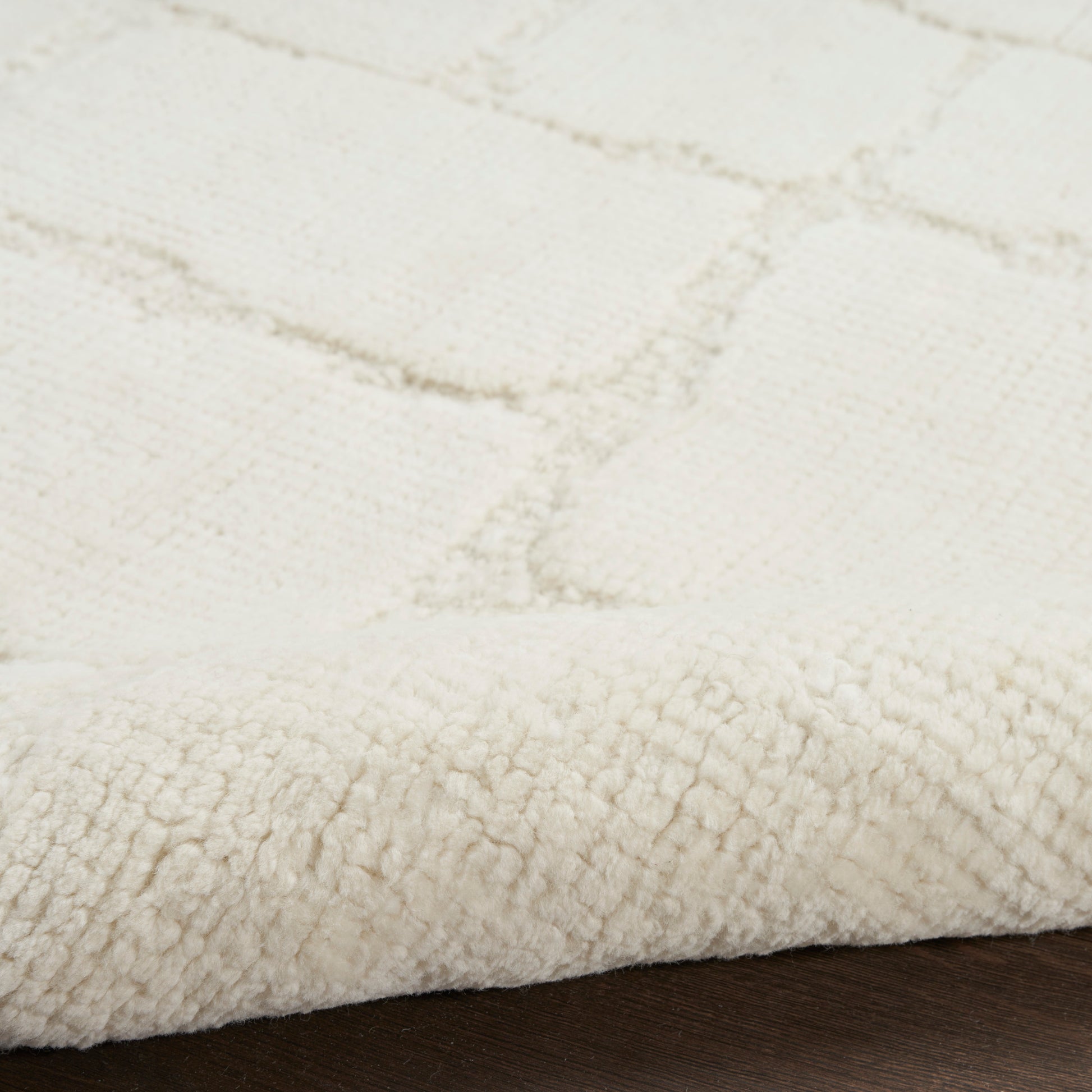 Cream plush rug close-up with thick pile and subtle grid pattern; edge folded on dark wood floor.