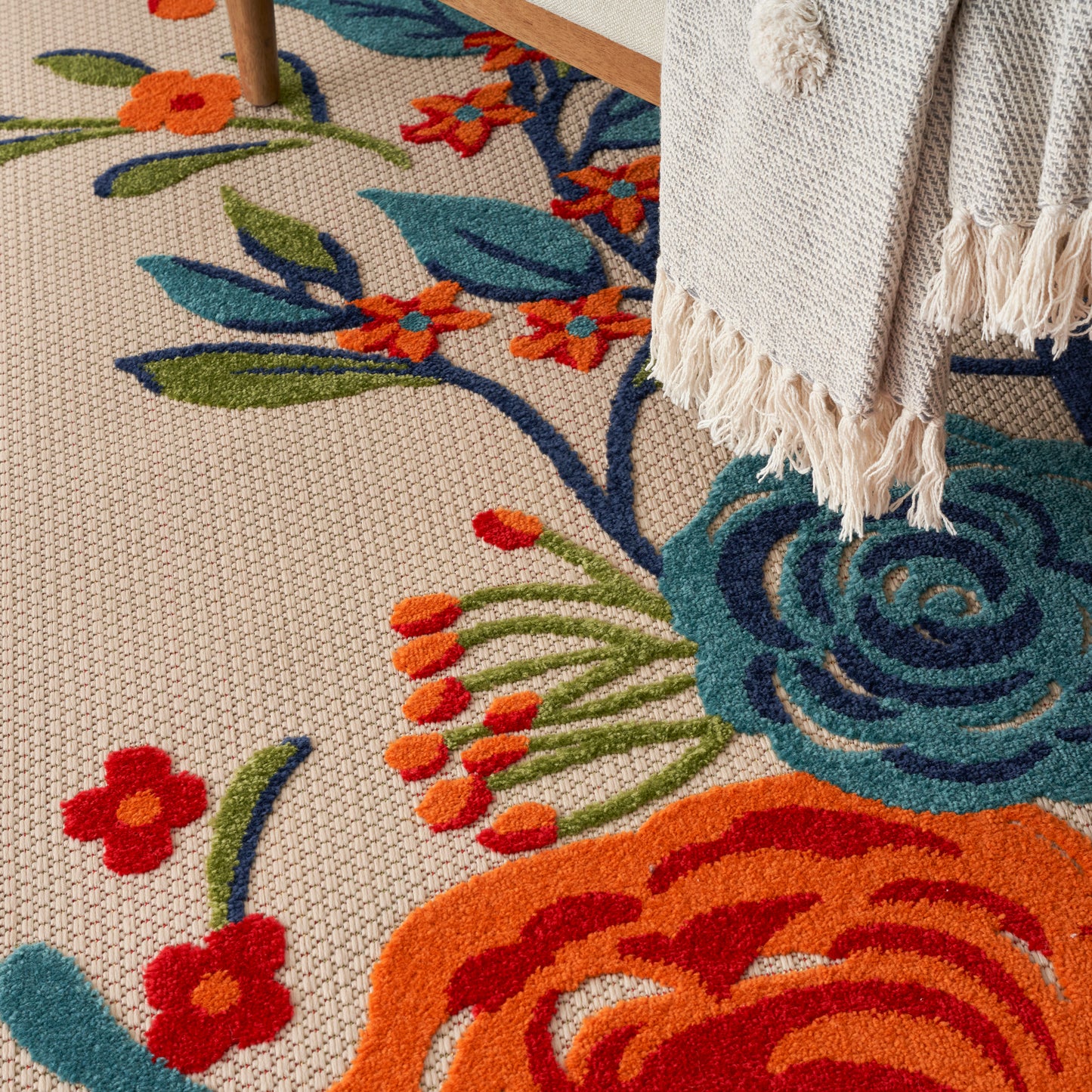 Colorful floral area rug with teal and orange blossoms on a beige woven base, plus a cream fringed throw draped over the edge.