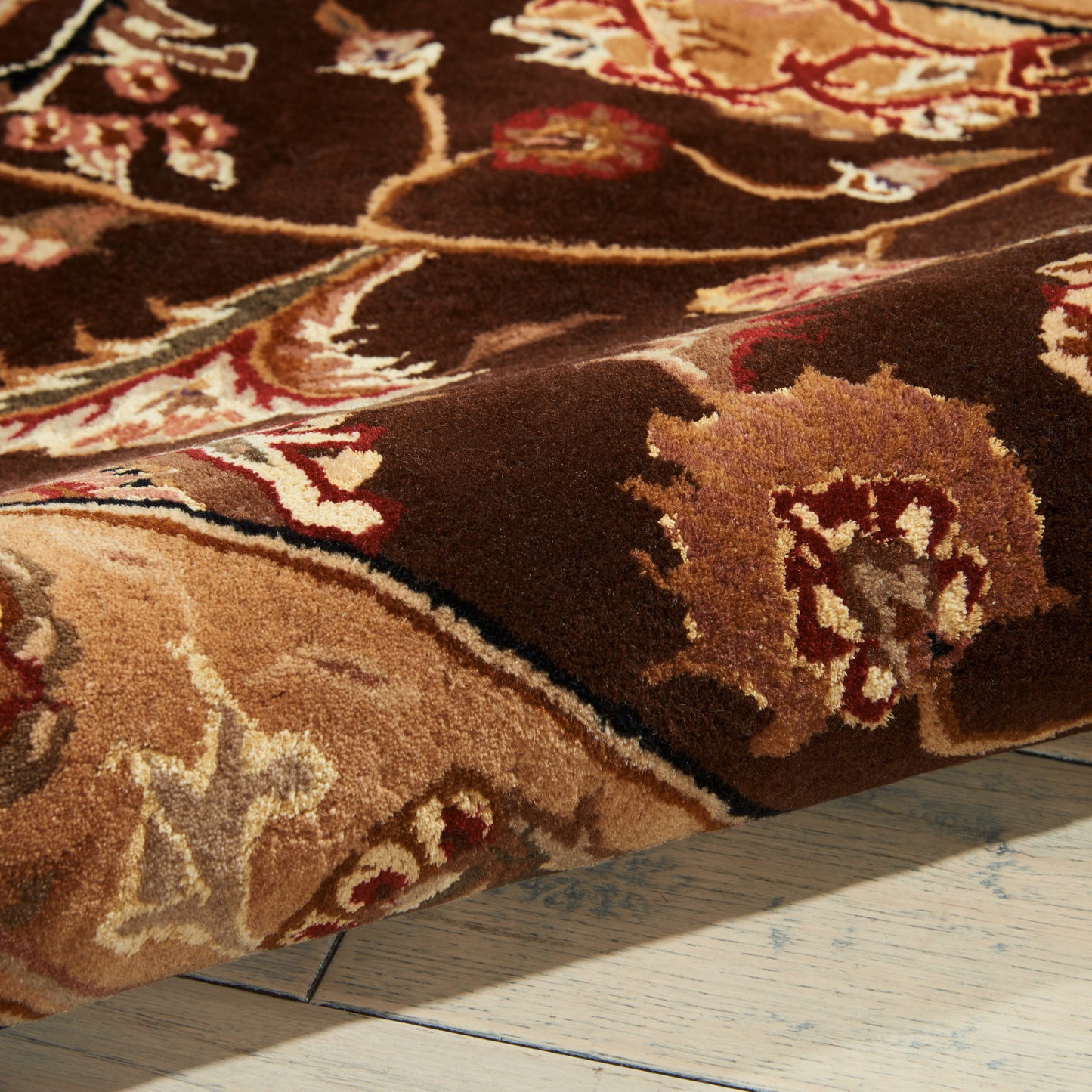 Close-up of a brown Persian-style rug with beige, tan, cream and red floral patterns, curled edge on a light wooden floor.