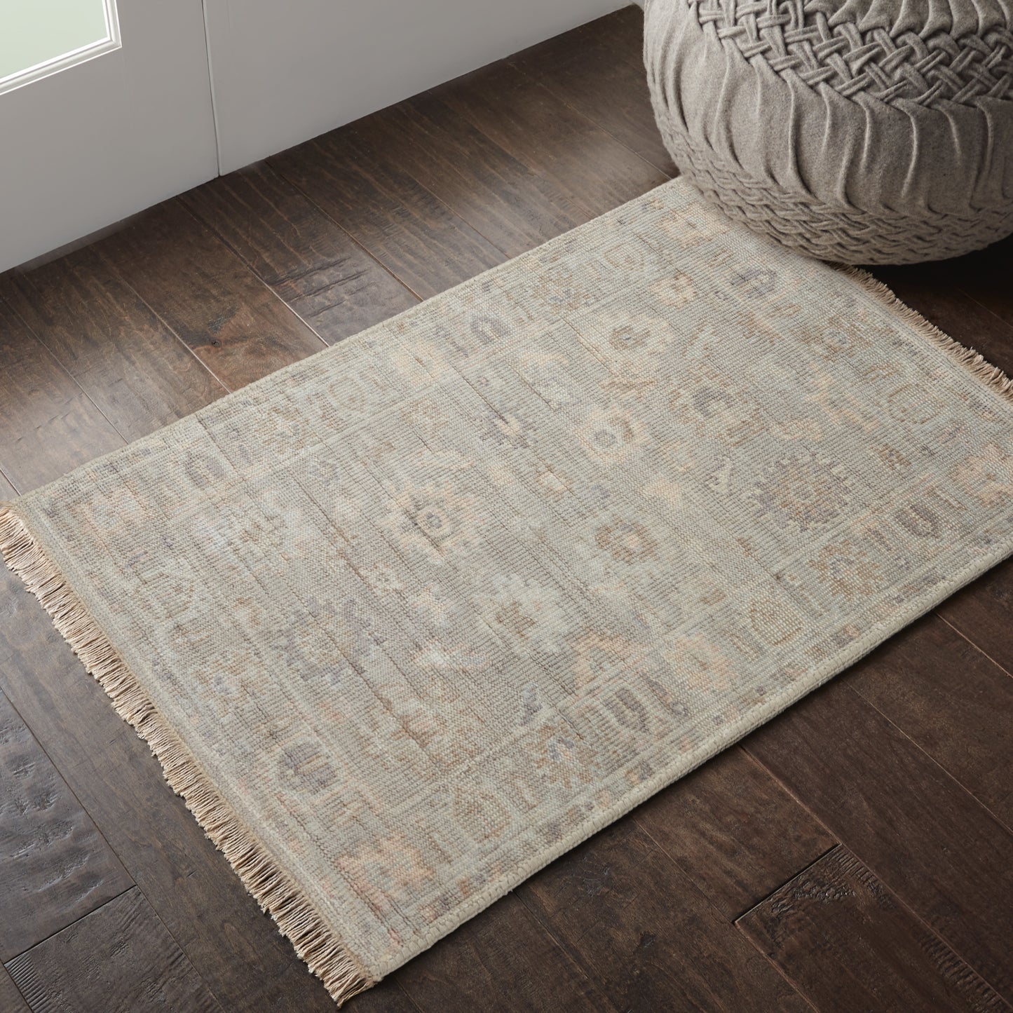 Beige flatweave rug with fringe, subtle geometric motif on dark wood floor beside a woven pouf.