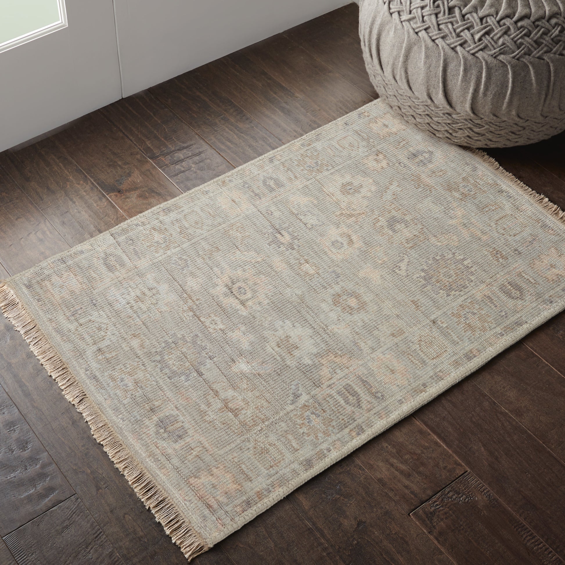 Beige flatweave rug with fringe, subtle geometric motif on dark wood floor beside a woven pouf.