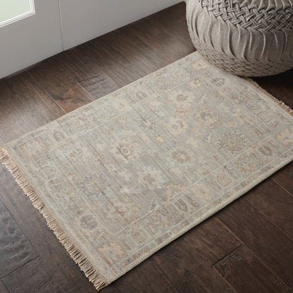 Beige flatweave rug with fringe, subtle geometric motif on dark wood floor beside a woven pouf.