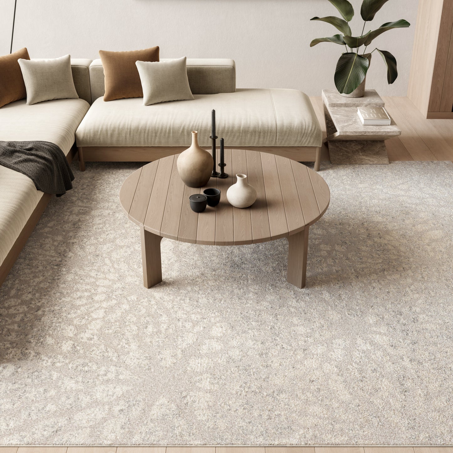 Beige sectional sofa with brown and beige pillows, round wooden coffee table with vases and cups, plant in background.