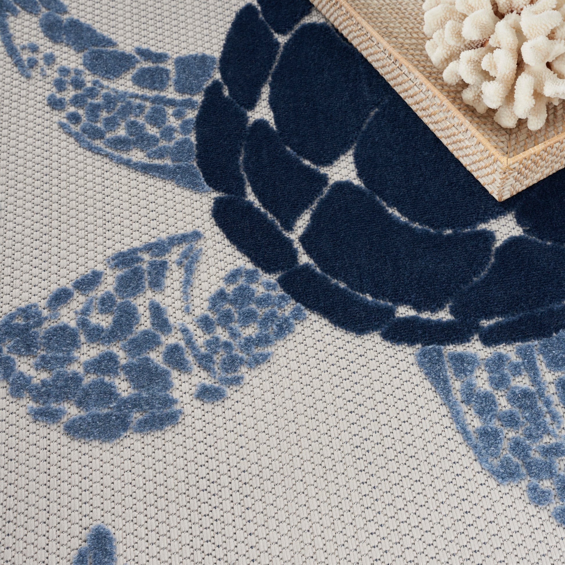 Blue floral rug with a large navy flower and pale blue petals on a beige textured background, braided mat corner.