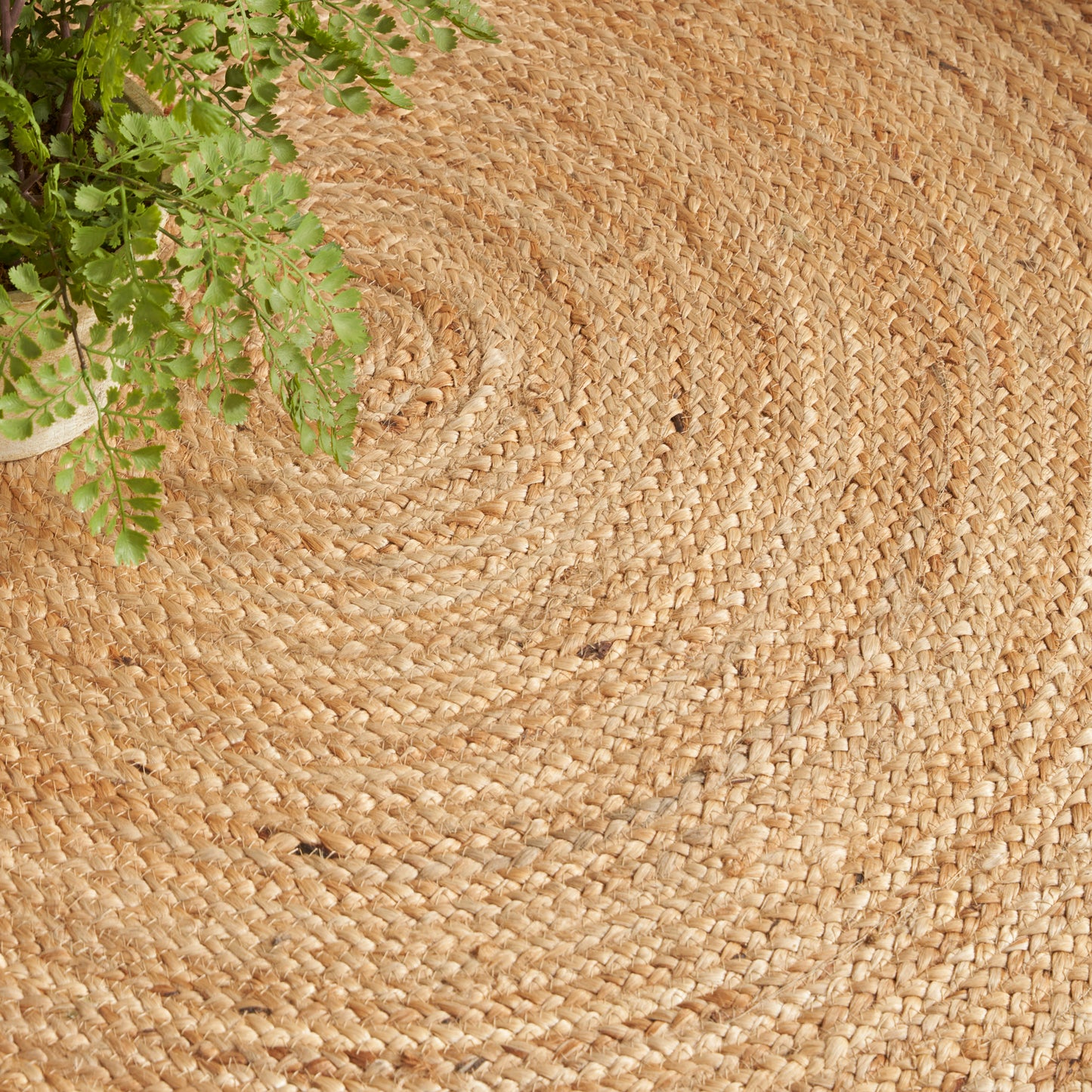 Round natural jute rug with concentric weave; green fern in the top-left corner, rustic boho decor.