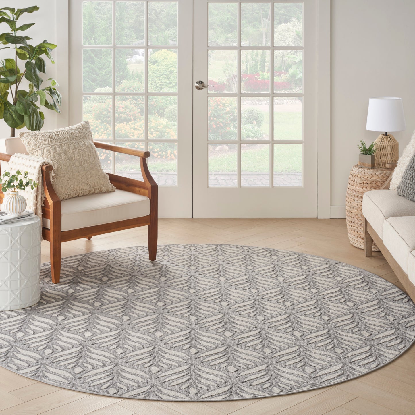 Round gray and ivory leaf-pattern area rug in a bright living room with wood chair, wicker side table, and French doors.