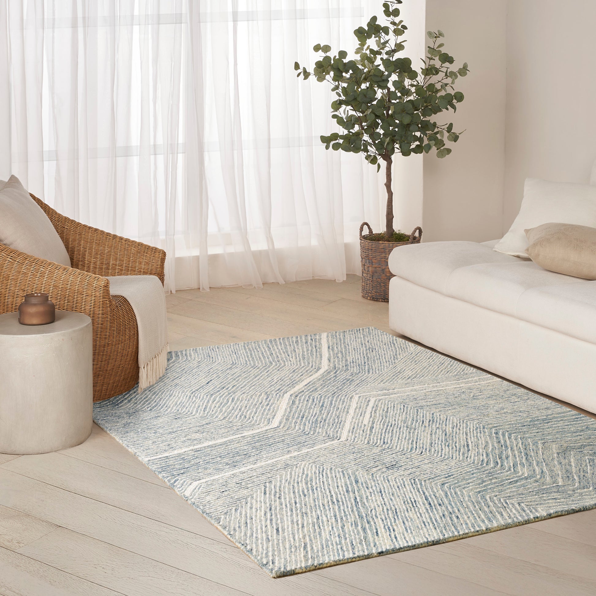 Bright living room with light wood floors, pale blue chevron rug, white sofa, wicker chair, plant and sheer curtains.