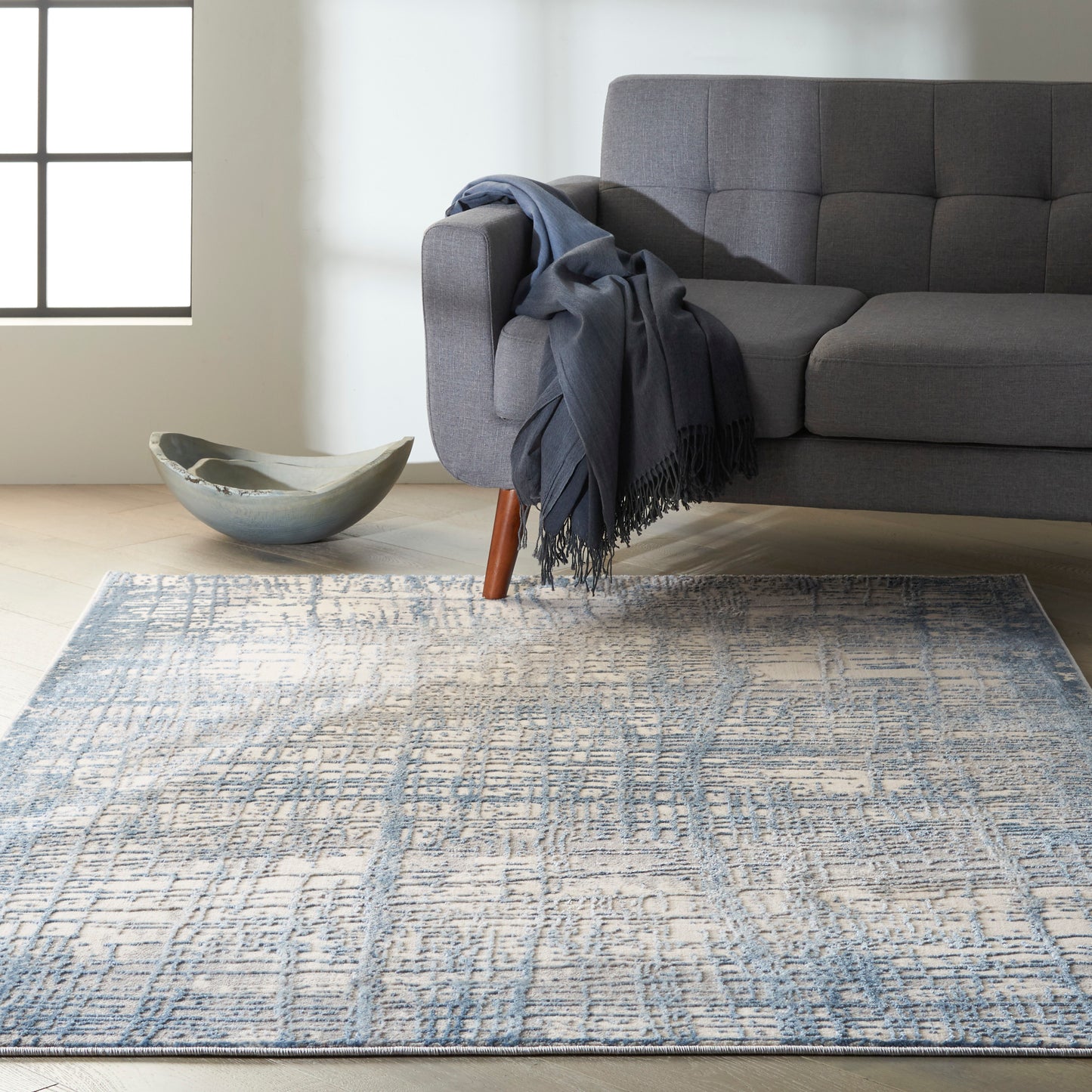 Grey upholstered sofa with wooden legs, blue throw blanket; ceramic bowl on light wood floor, blue-toned geometric area rug.