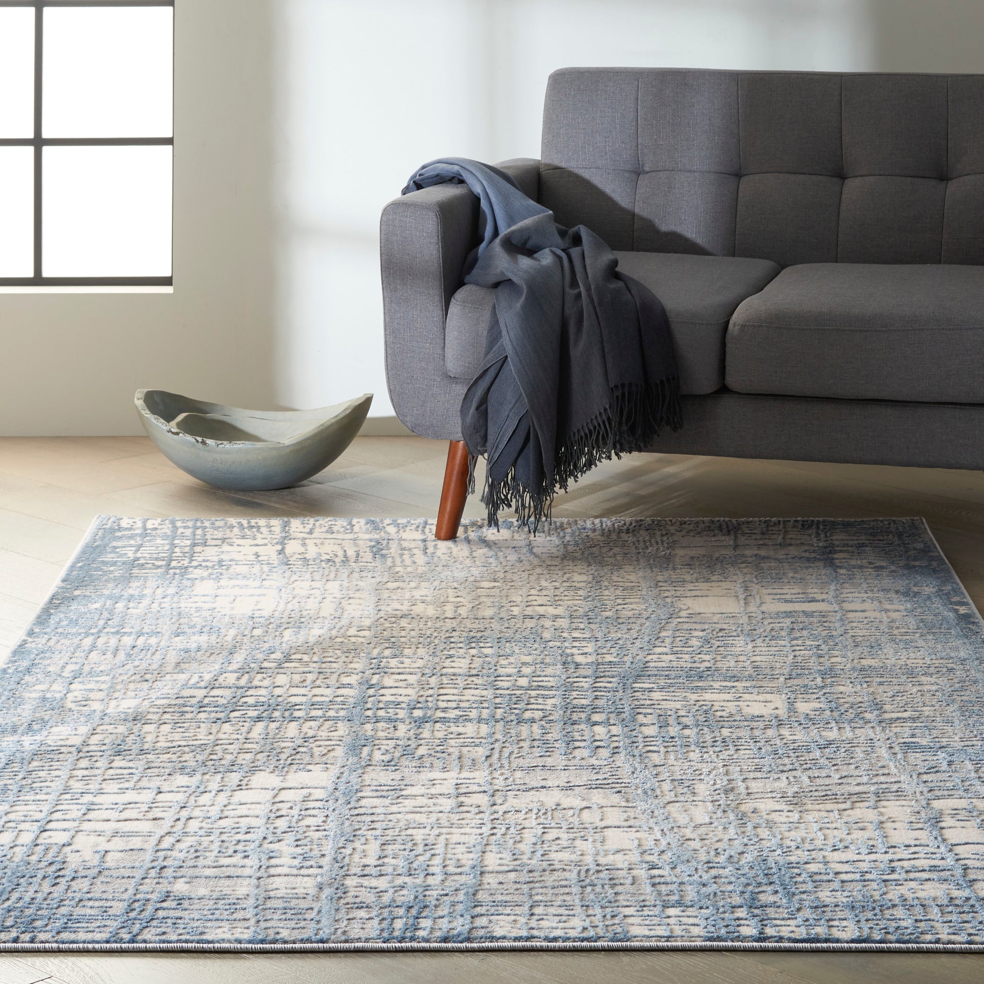 Grey upholstered sofa with wooden legs, blue throw blanket; ceramic bowl on light wood floor, blue-toned geometric area rug.