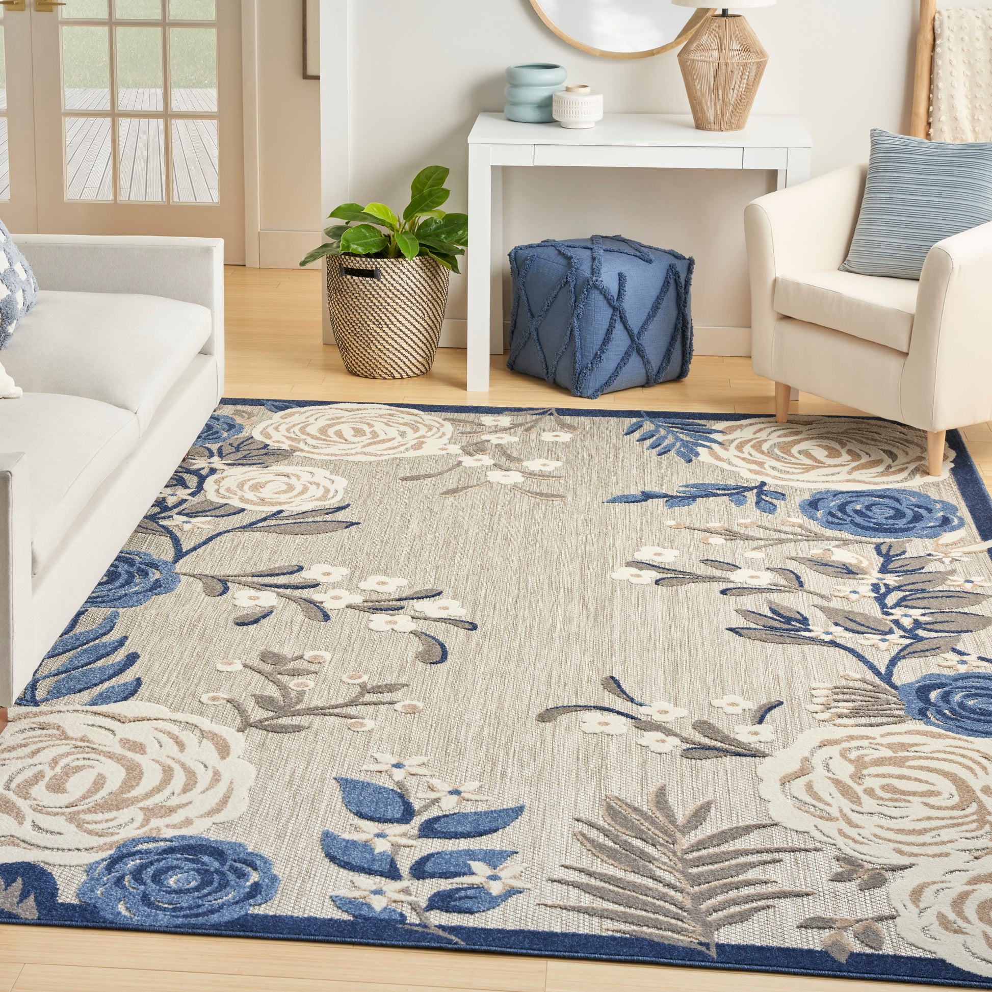 Floral area rug in blues, beiges, and grays on a light wood floor, with cream sofa, plant, blue pouf, and white console.