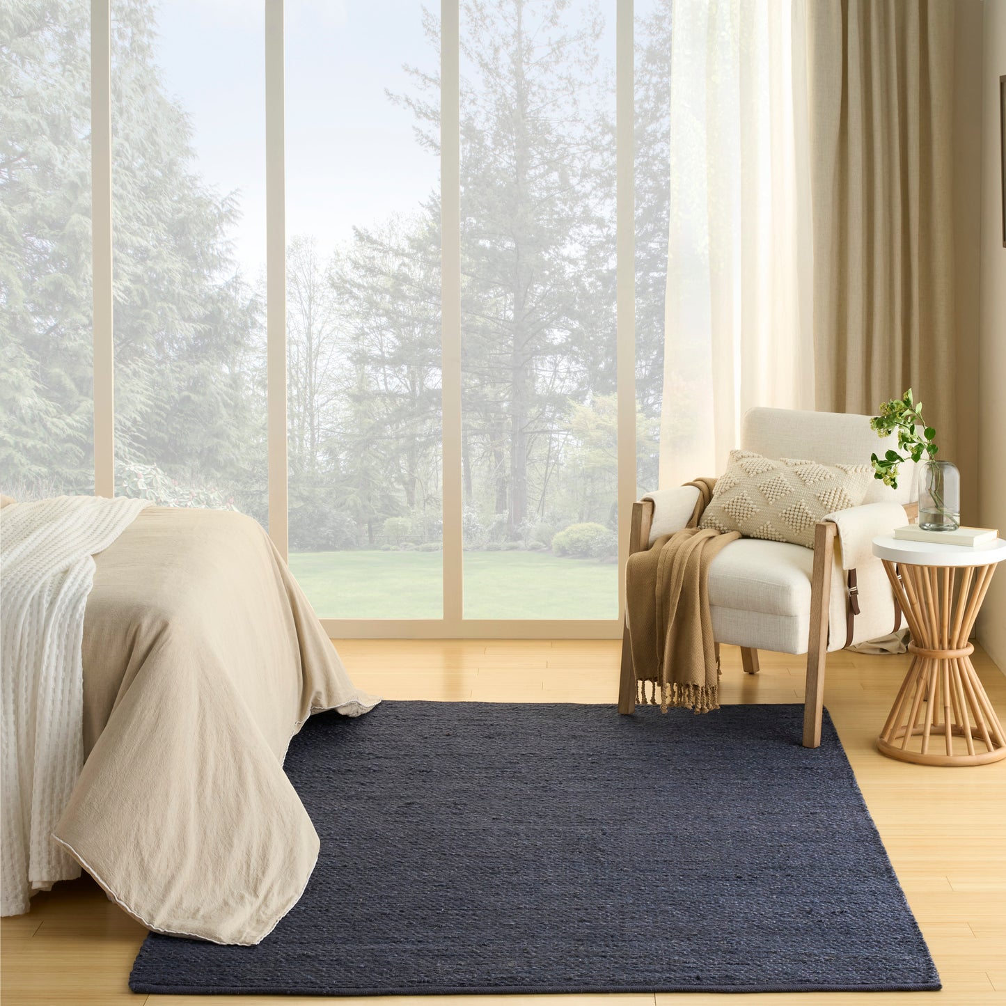 Cozy bedroom: beige bed, navy rug, light wood floor, white armchair with beige throw, round side table with plant, windows.