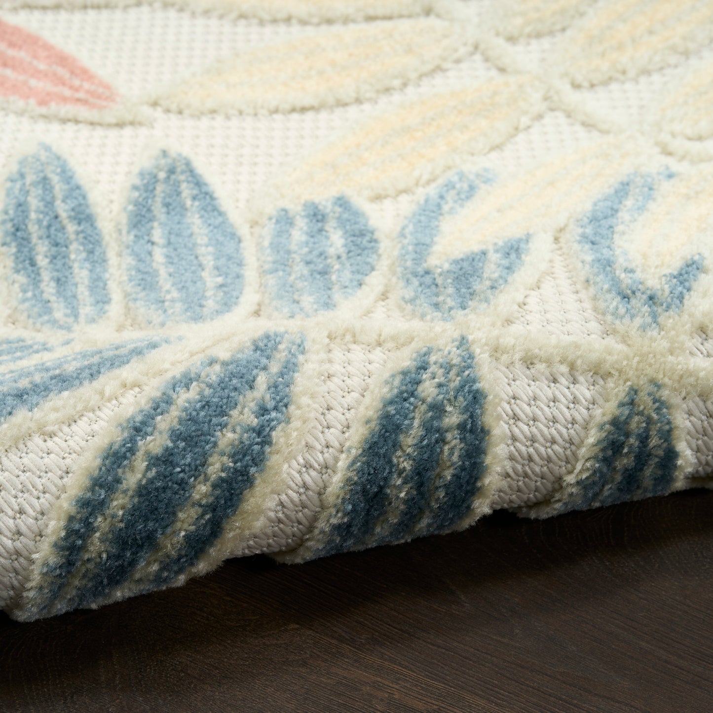 Cream rug with blue leaf-pattern, plush pile texture, folded edge showing thick weave, on dark wood floor.