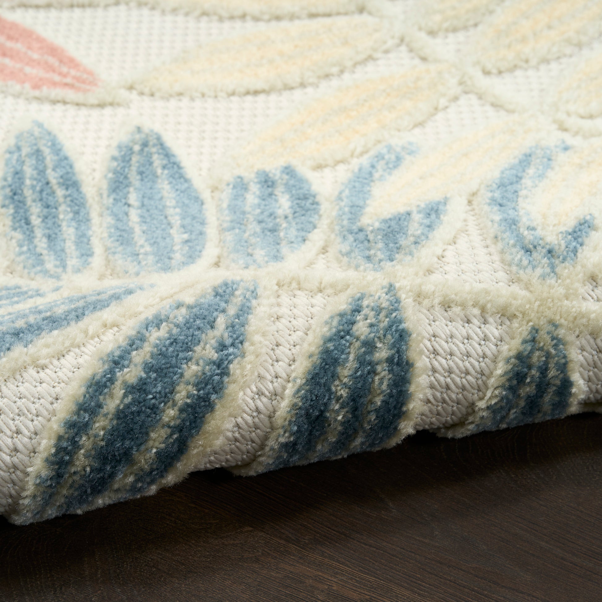 Cream rug with blue leaf-pattern, plush pile texture, folded edge showing thick weave, on dark wood floor.