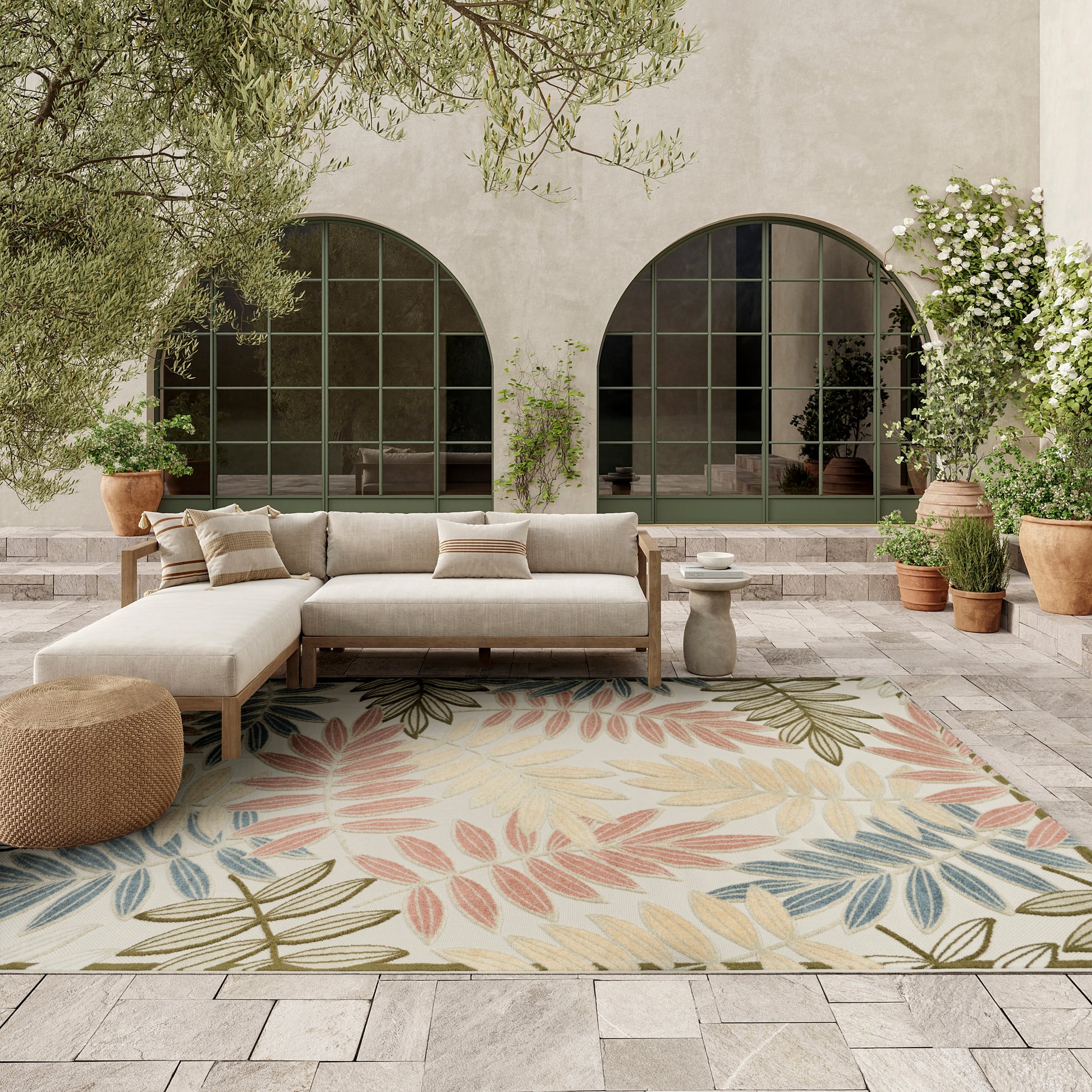 Outdoor patio with beige sectional sofa, pastel leaf rug, arched windows, & potted plants.