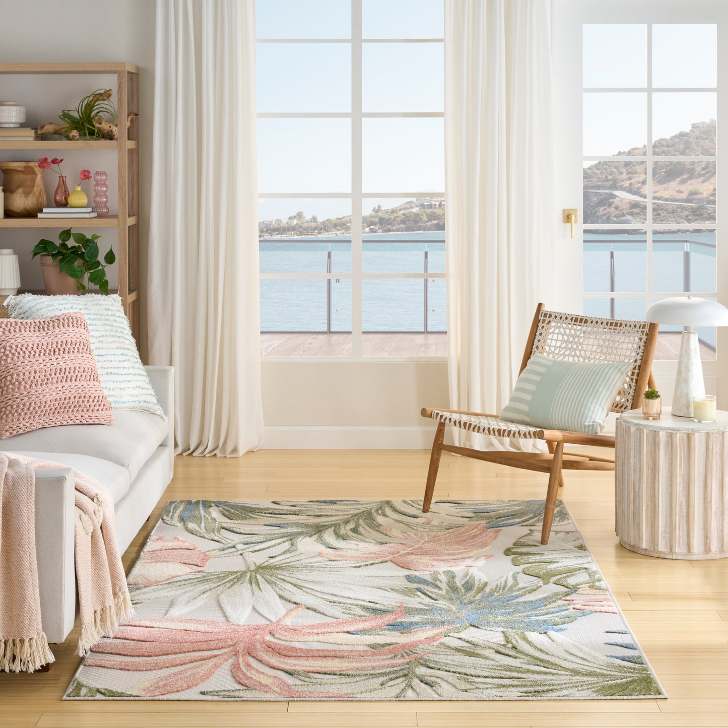 Coastal living room with large ocean-view window, pastel tropical rug, light sofa with pink & mint cushions, woven chair & wood side table.