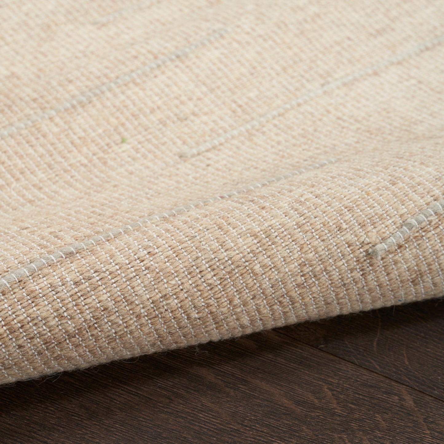 Beige woven rug with subtle white stripes and folded edge on dark wood floor.