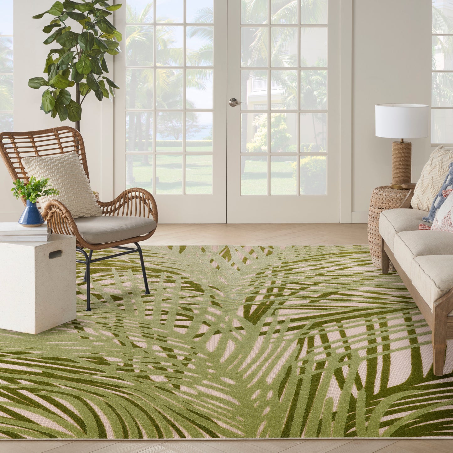 Living room with palm-leaf area rug in olive greens, wicker chair, neutral sofa, potted plant, glass-pane doors.