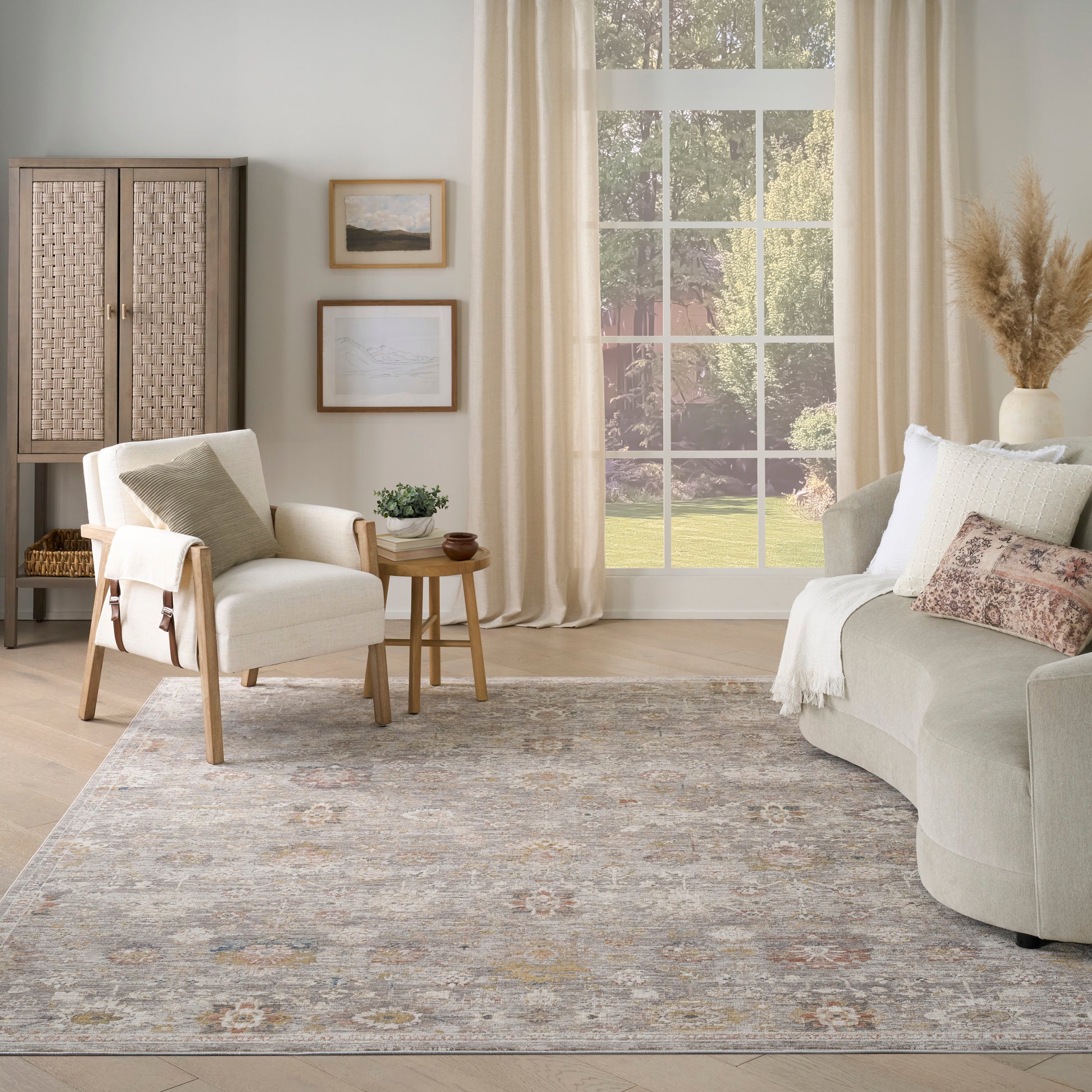 Neutral living room with cream sofa, beige chairs, round wood table, woven cabinet, framed art, large window, gray patterned rug, dried grasses