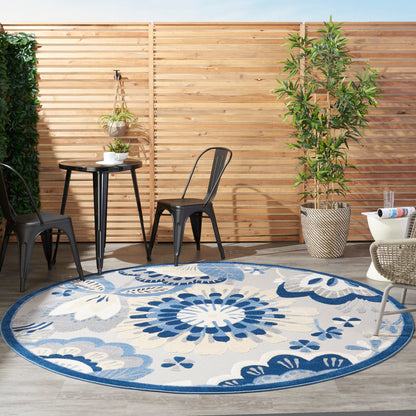 Round blue & white floral outdoor rug on wooden deck with black metal chairs, round table, hanging planter & potted tree.