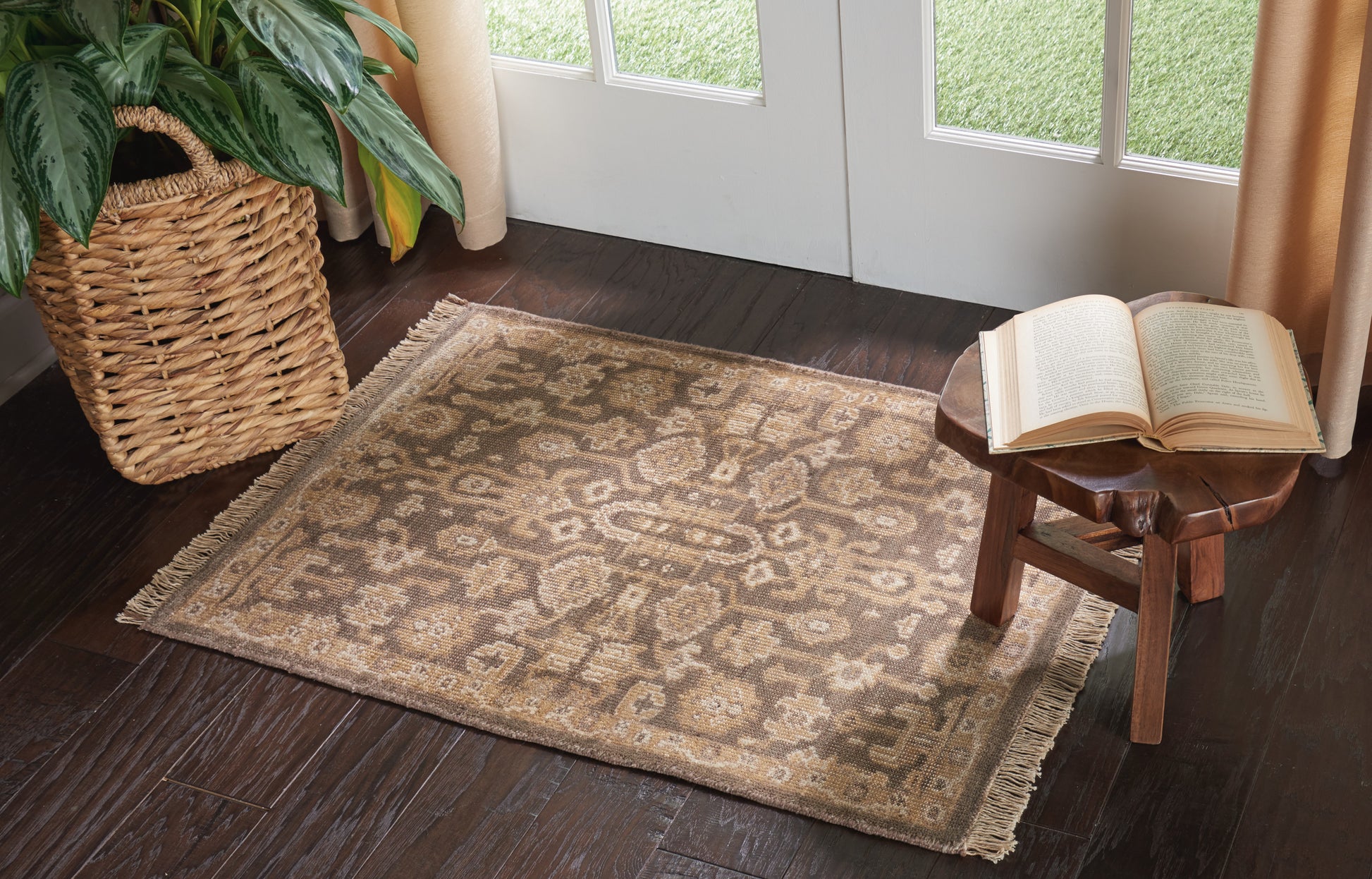Brown-toned oriental rug with fringed edges on dark wood floor, rustic wooden stool and open book, woven plant basket nearby.