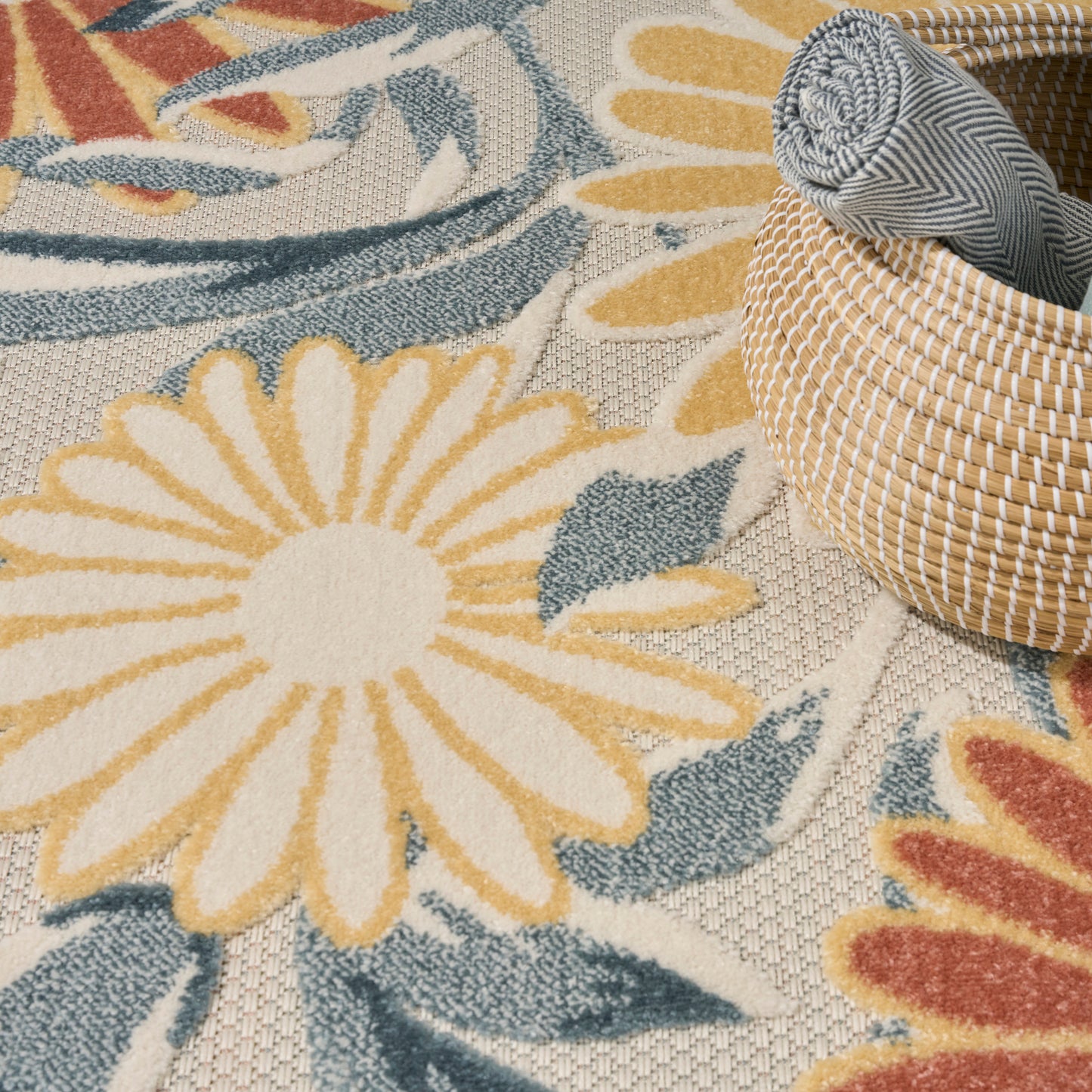 Textured floral rug with cream-yellow daisies, blue-green leaves; round wicker basket and rolled striped fabric.