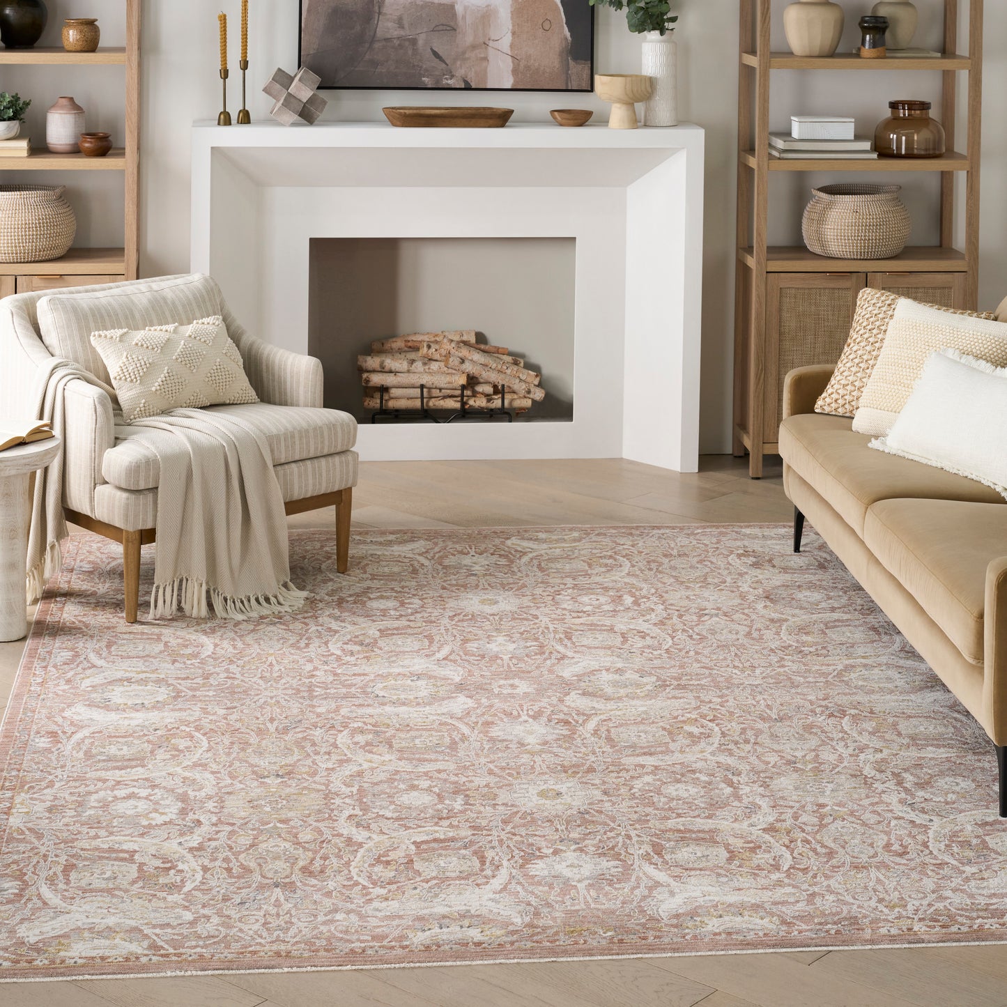 Cozy beige living room with plush cream armchair, tan sofa, ornate pinkish area rug, white fireplace, and wooden shelving.