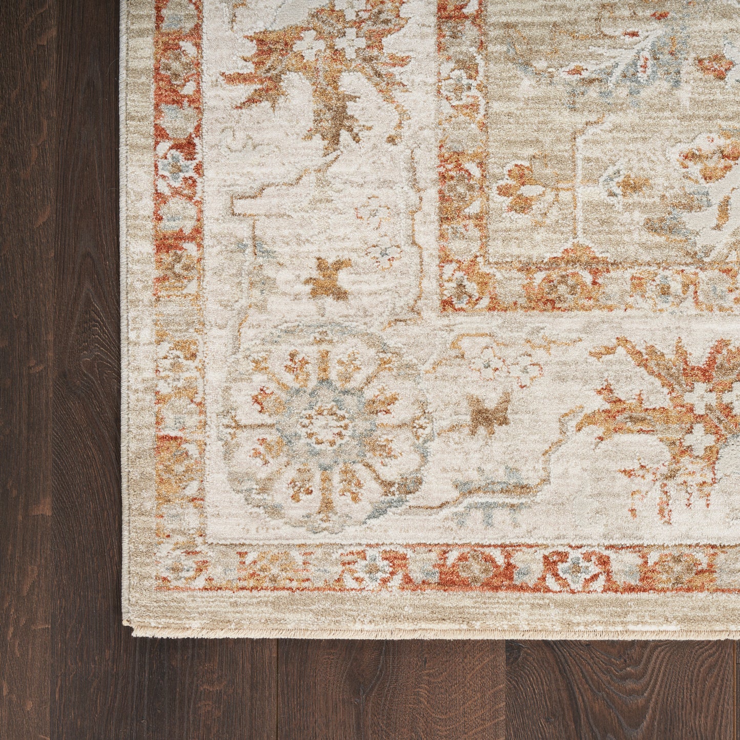 Beige oriental rug with rust and sky blue floral medallion border on dark wood floor, fringe at edge.