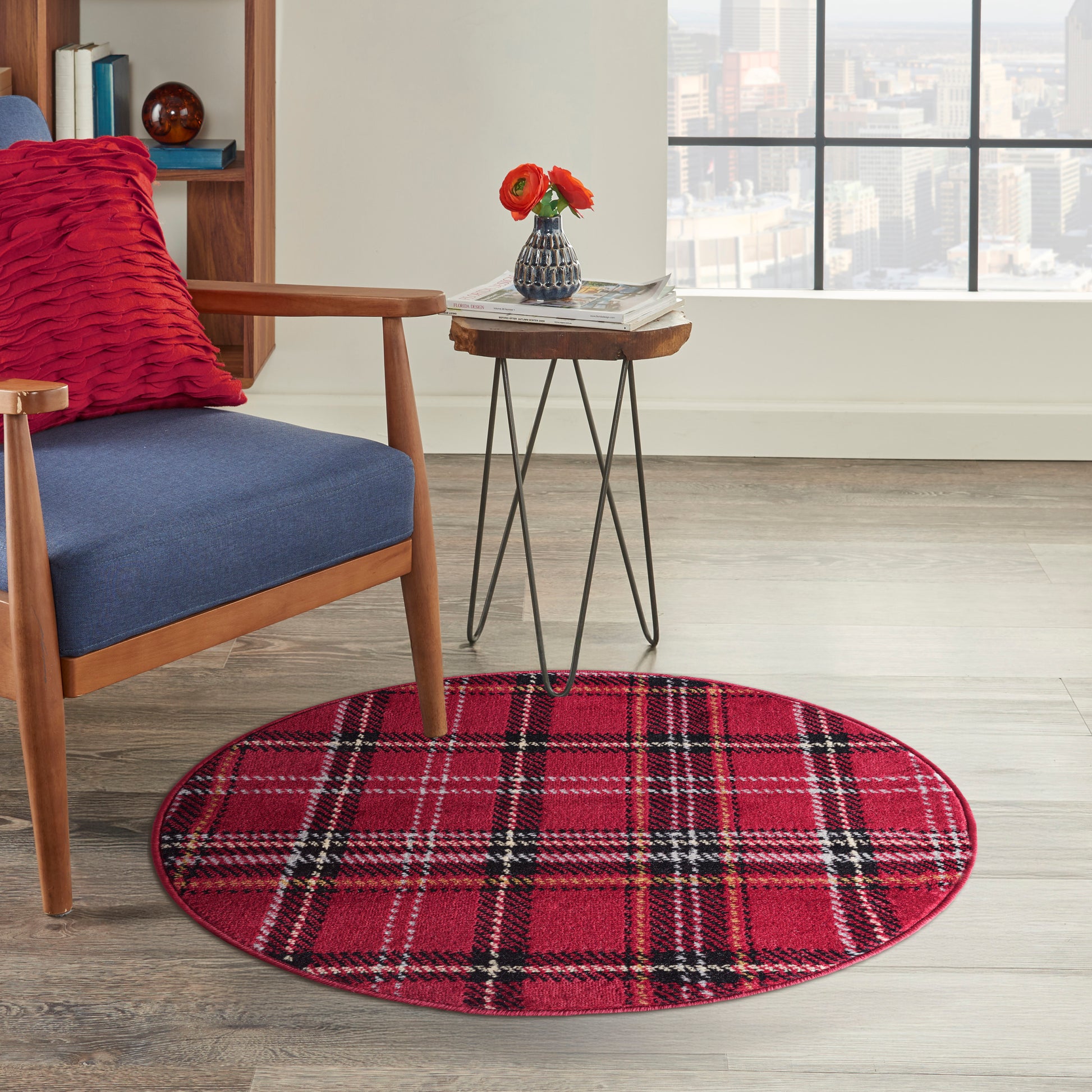 Circular red plaid rug under a blue upholstered mid-century chair, side table with magazines and a vase of red flowers.