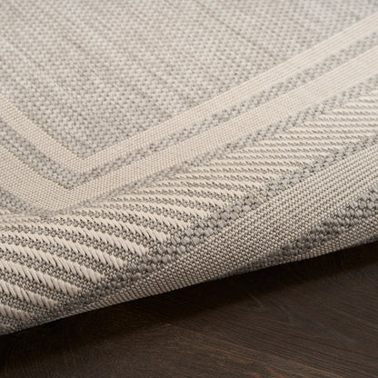 Close-up of a beige-gray woven rug with a bordered geometric pattern and a folded corner on dark wood floor.