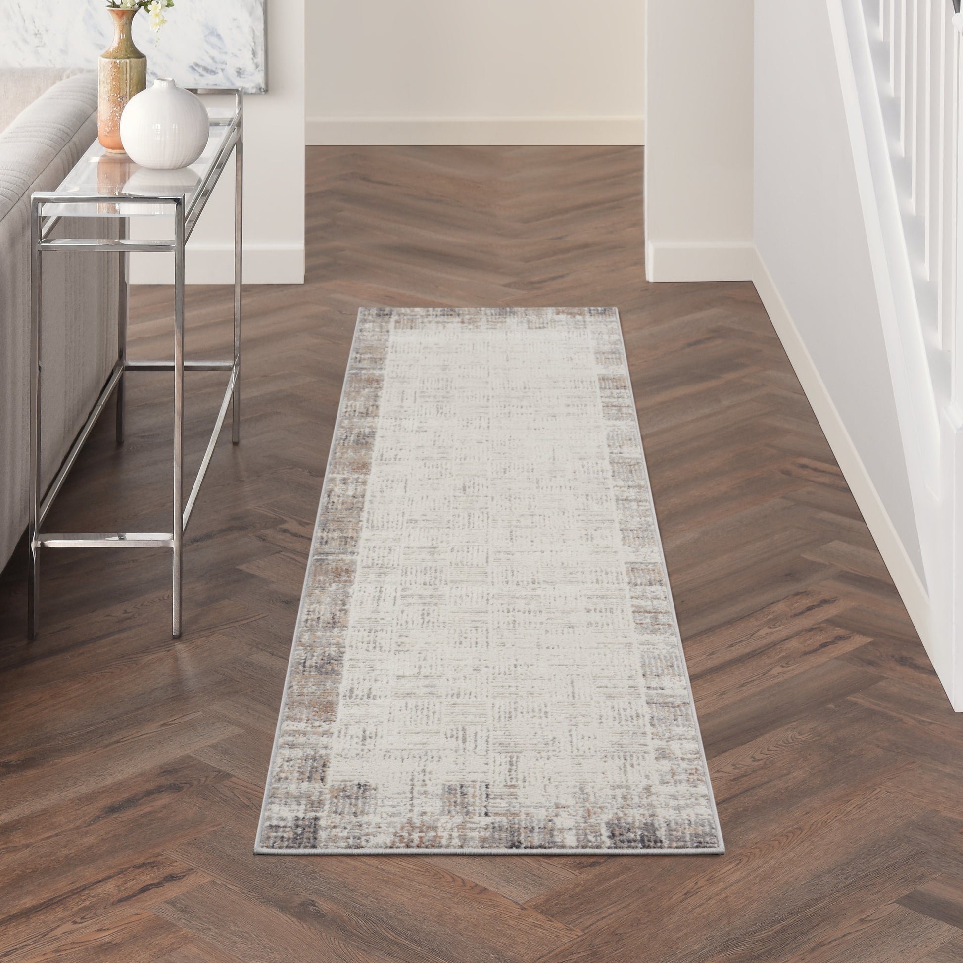 Long neutral-toned runner rug with a subtle pattern on a dark herringbone hallway floor; glass console table on the left.