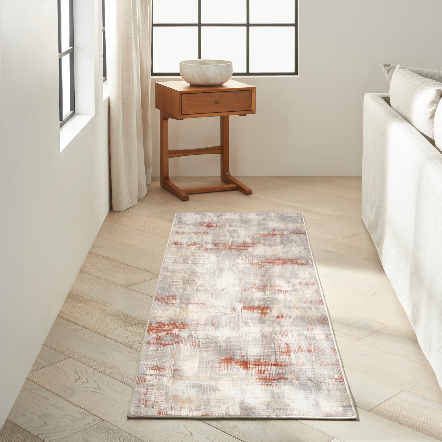 Abstract runner rug in cream, gray and rust tones on light wood floor beside a wooden side table and cream sofa.