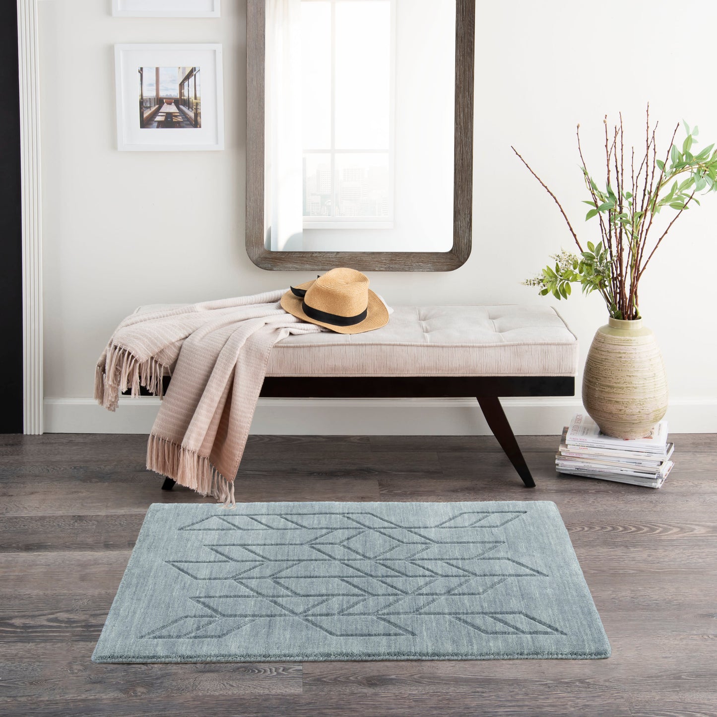 Beige tufted bench with dark wood legs, beige throw and straw hat, mirrored wall, vase with branches, geometric rug.