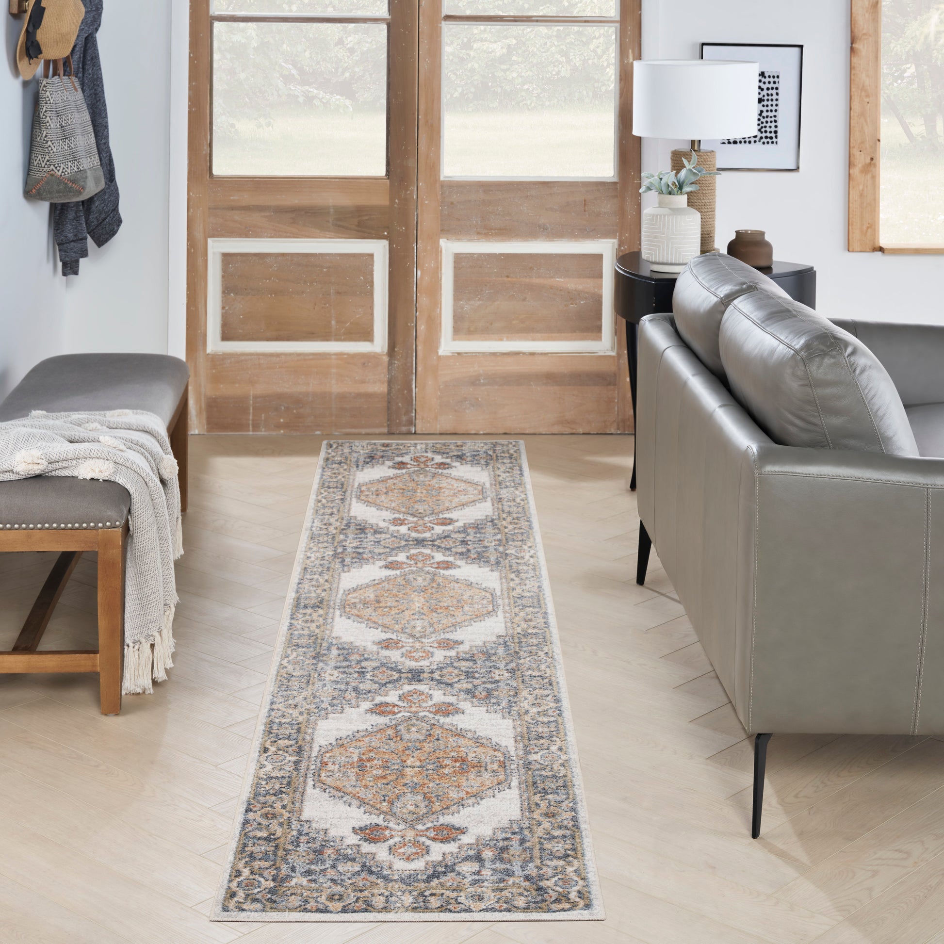 Long runner rug with geometric medallions in ivory, blue, rust on light wood floor beside gray sofa and wooden doors.