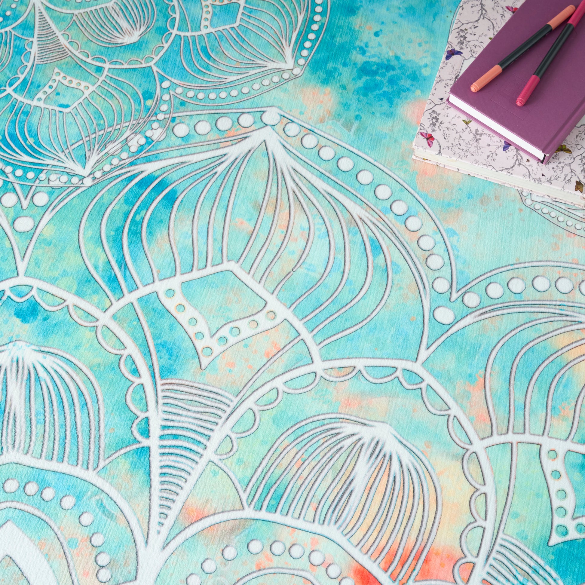 Turquoise tie-dye round rug with white mandala outlines; purple floral notebook and two colored pens in the top-right.