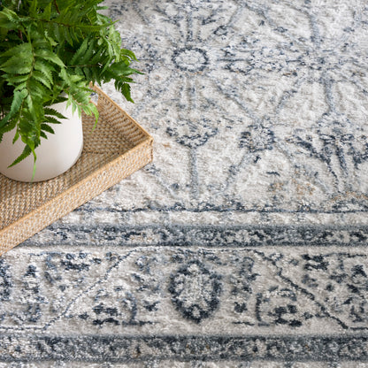 White ceramic pot with green fern on a woven rattan tray, resting on a grey-blue ornate patterned rug.